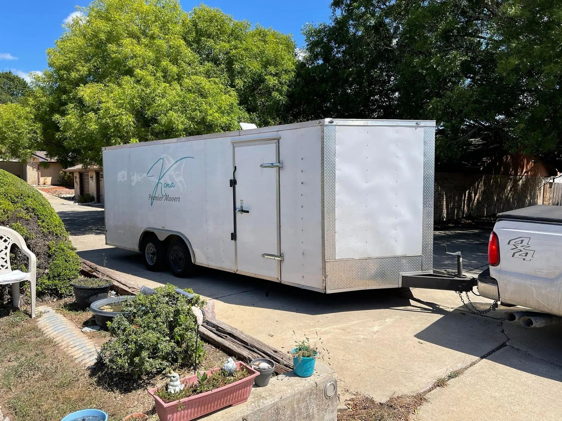 White enclosed trailer parked in a driveway beside a silver pickup, with trees and a house in the background