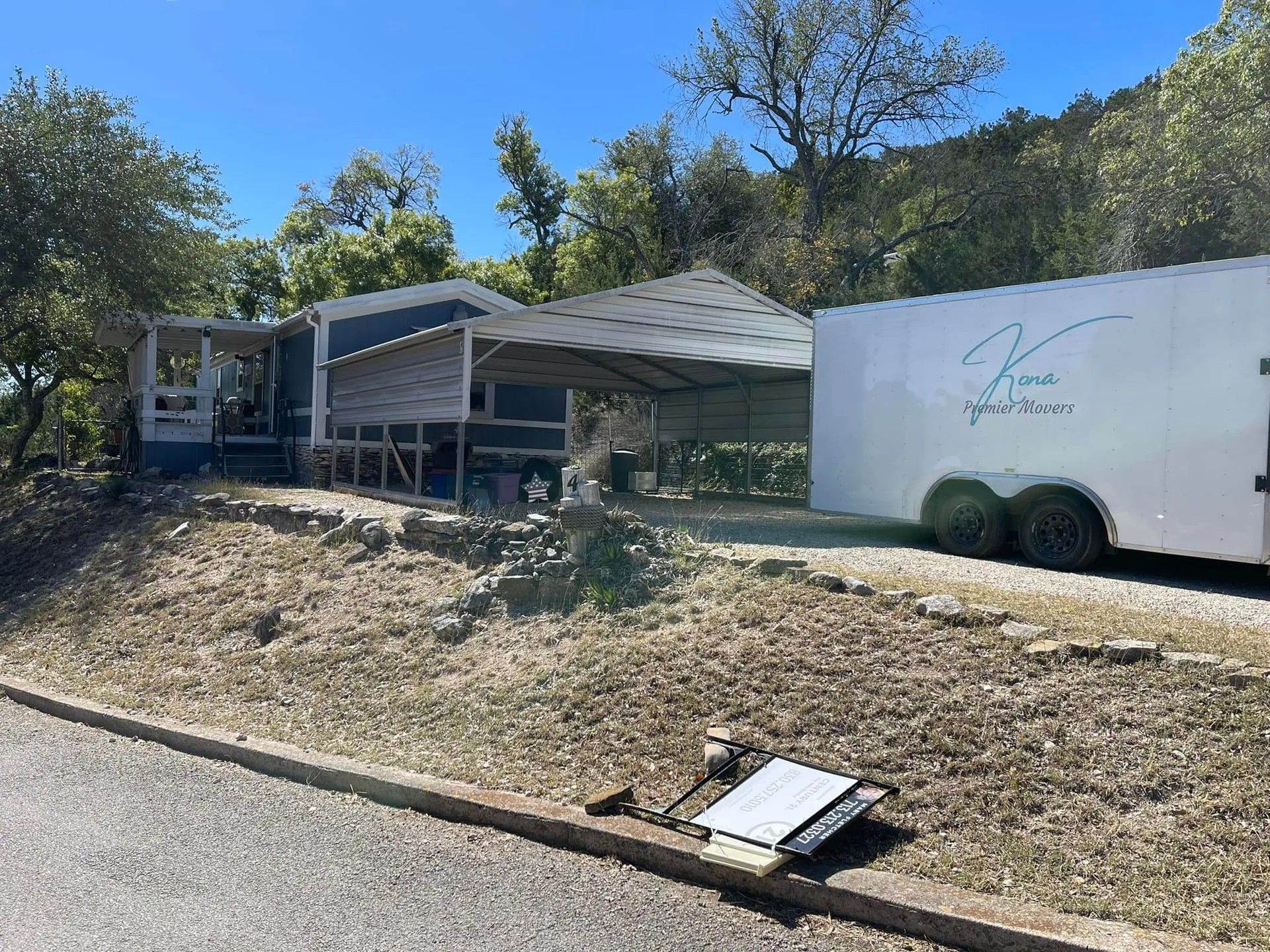Damaged roadside structure with metal roof and trailer beside a dry embankment under trees