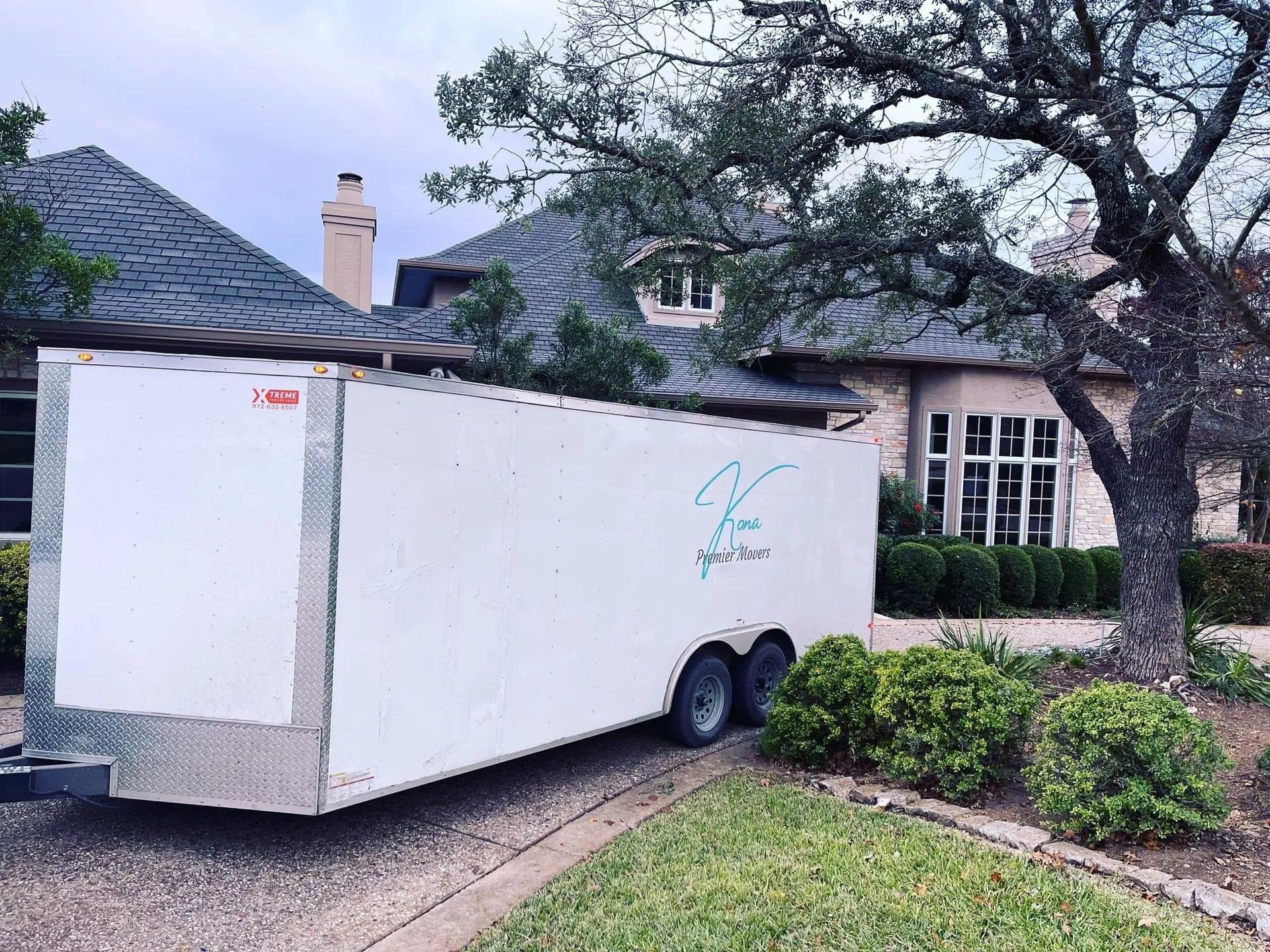 White cargo trailer parked in a driveway beside a house and tree, with shrubs in front