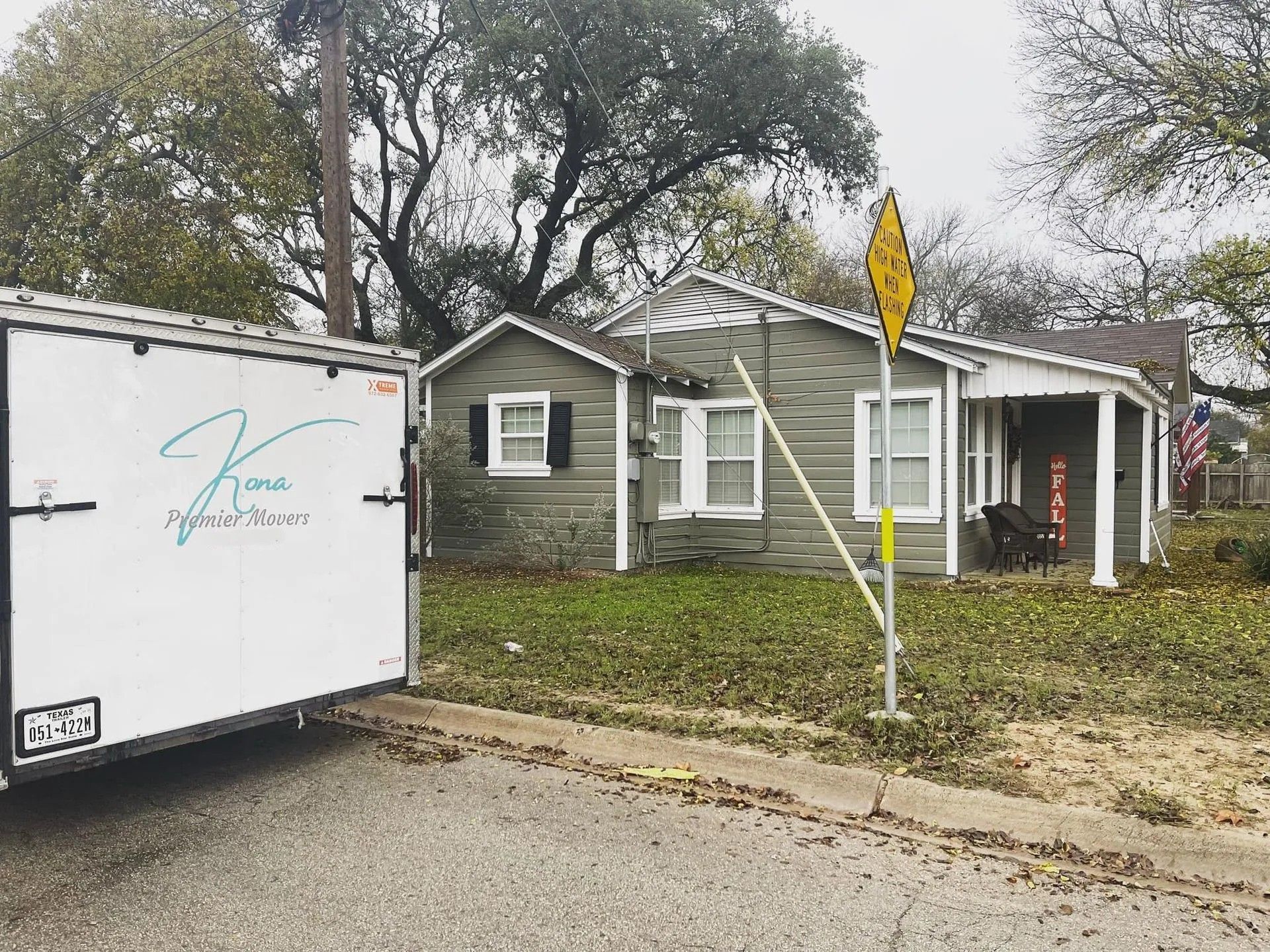 White trailer beside a small gray house on a leafy street, with a yellow road sign in front