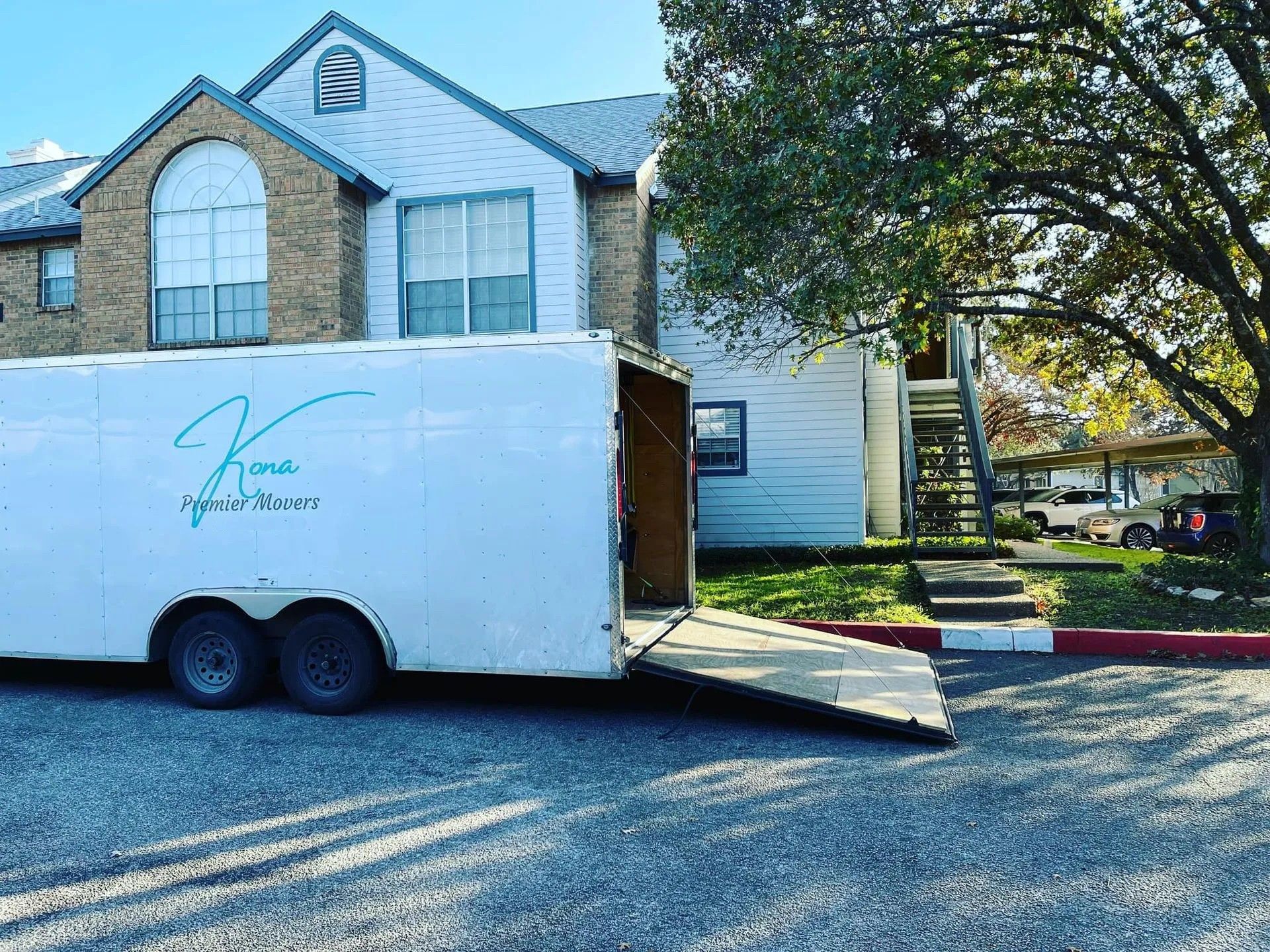 White trailer parked beside a suburban house with a ramp and open door, under a large tree.