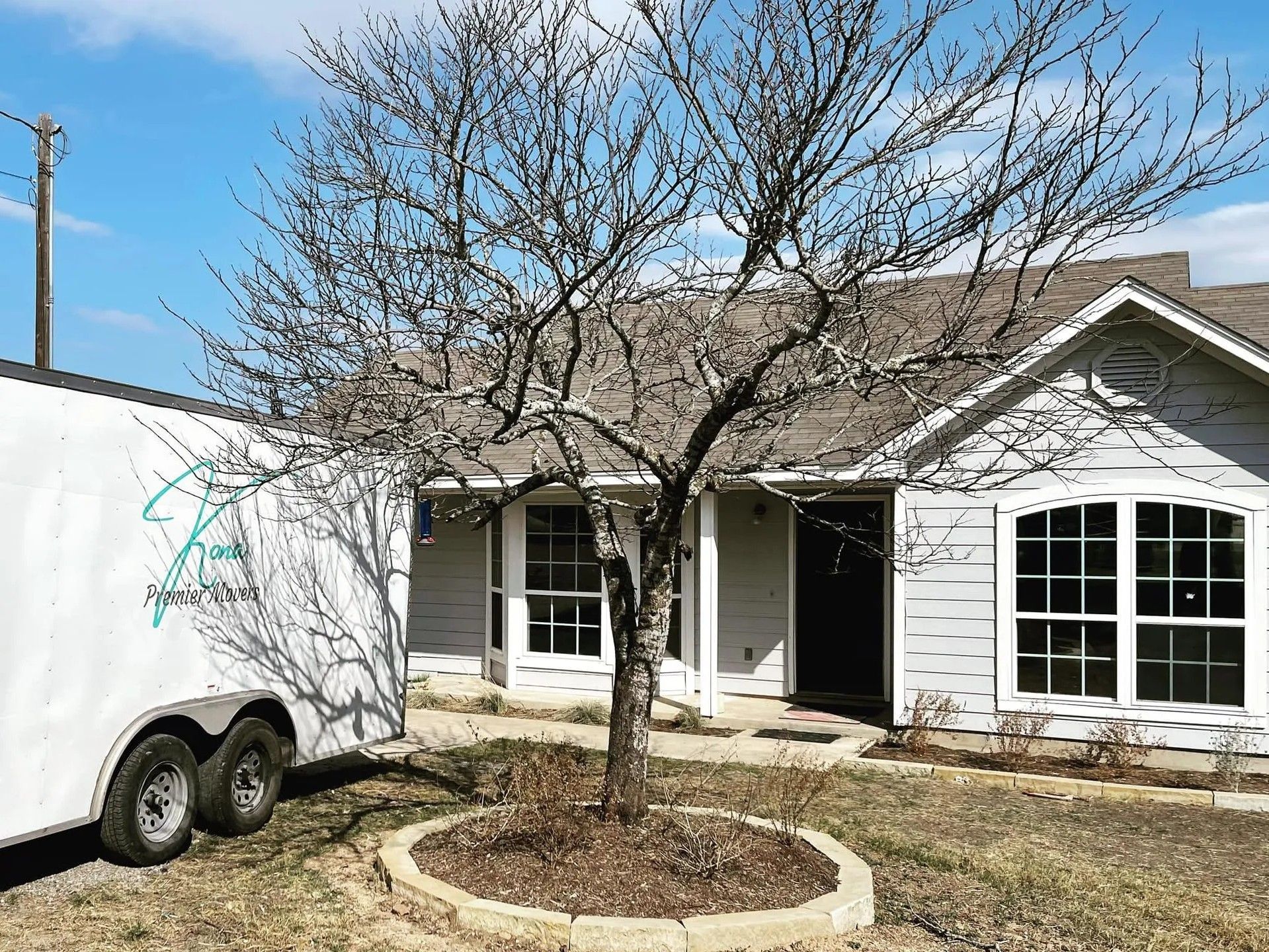 Single-story white house with large windows and a trailer in the front yard under a leafless tree