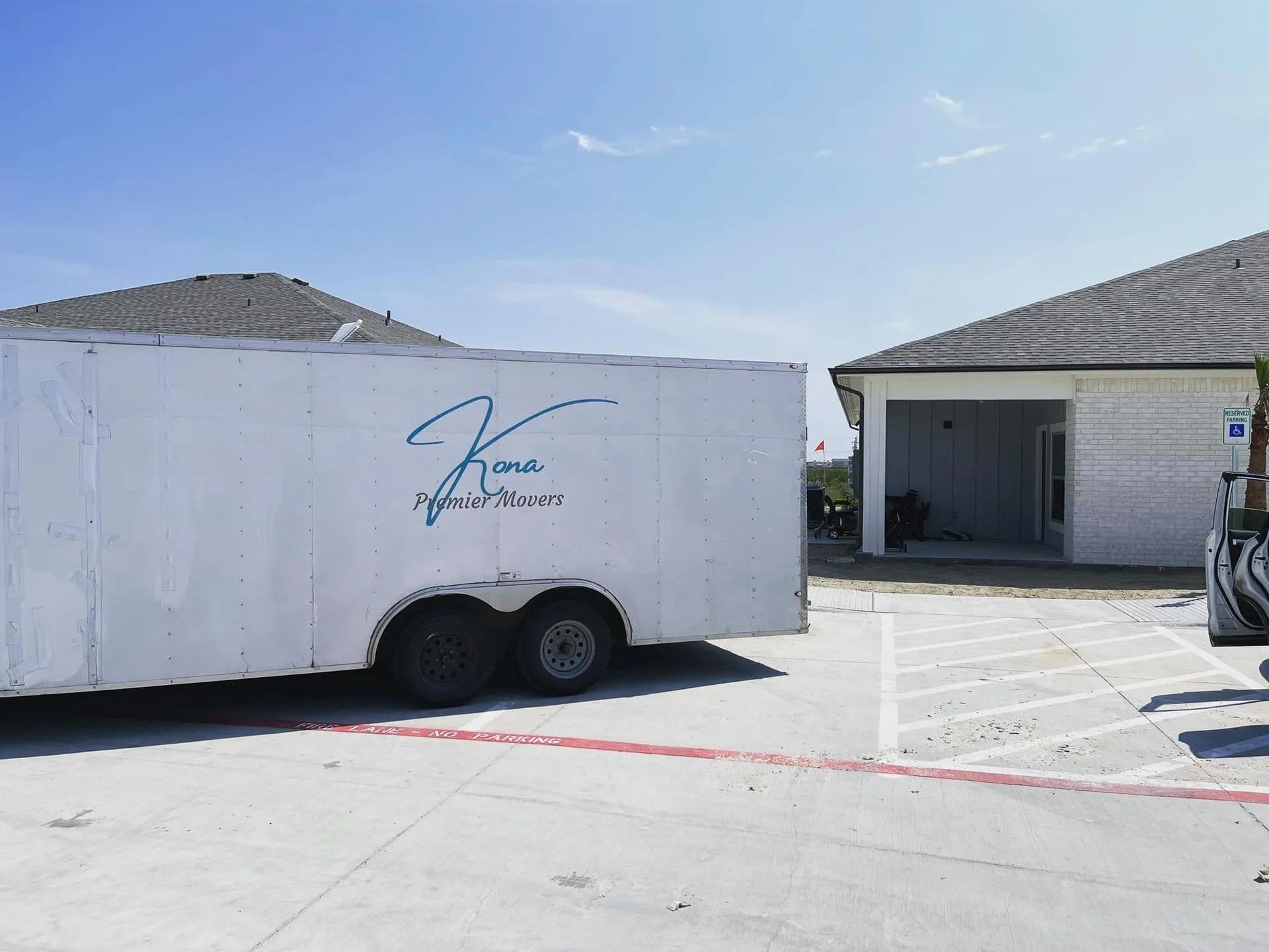 White utility trailer parked beside a building in a sunny lot, with another truck at the right edge.
