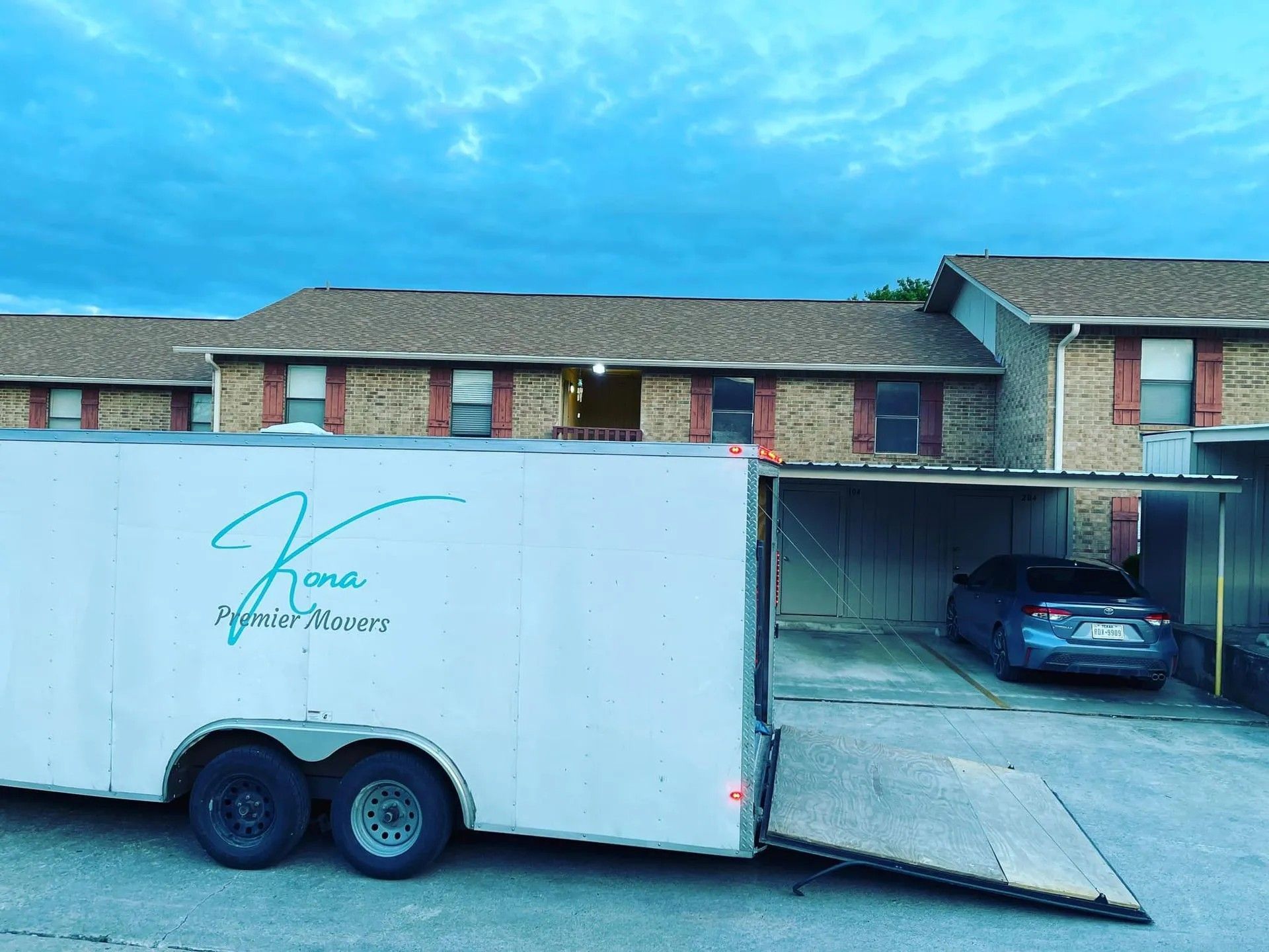 White enclosed trailer loaded with a blue car, parked before a brick building with its ramp lowered.