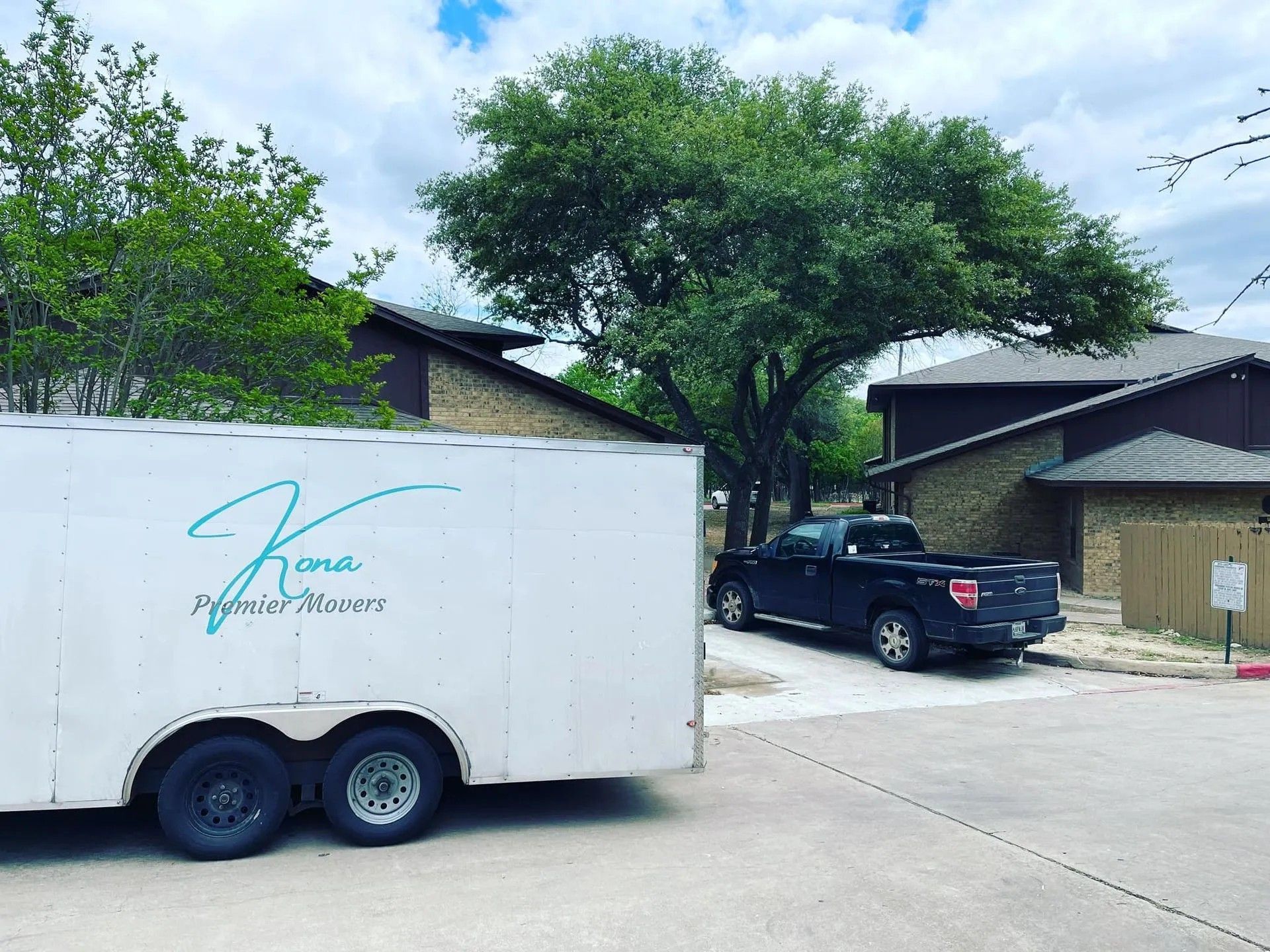 White dual-axle trailer on a driveway beside a black pickup and suburban houses under trees