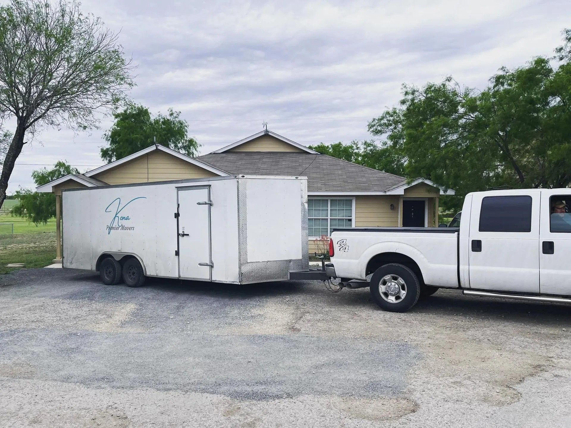 White pickup truck hitched to a white enclosed trailer in front of a house, on a gravel driveway.