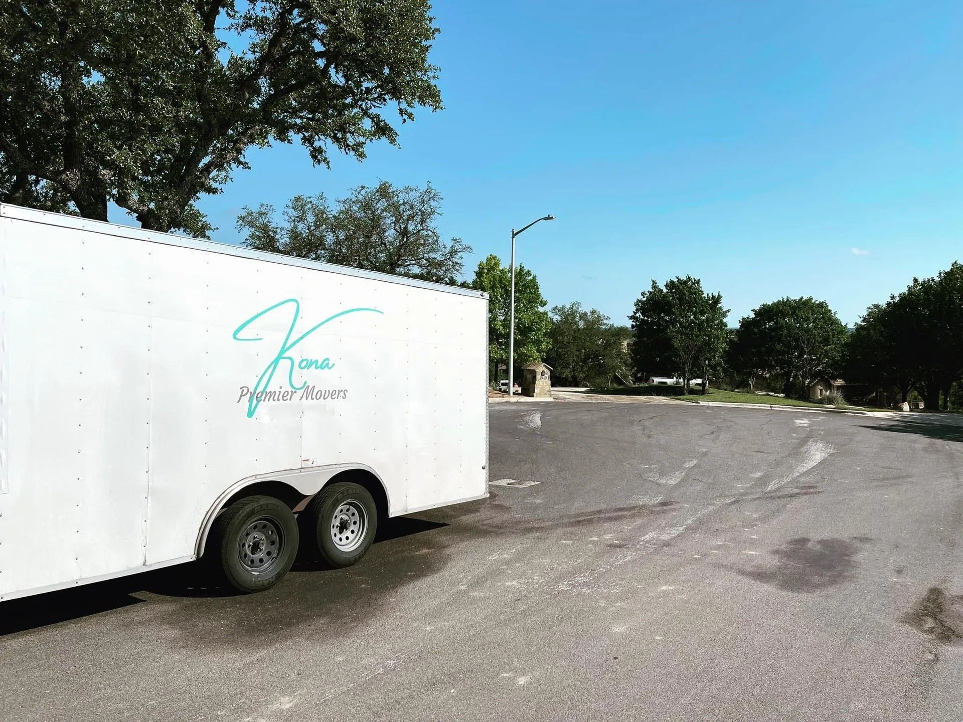 White utility trailer parked on a sunny road beside trees and a curve in the pavement.
