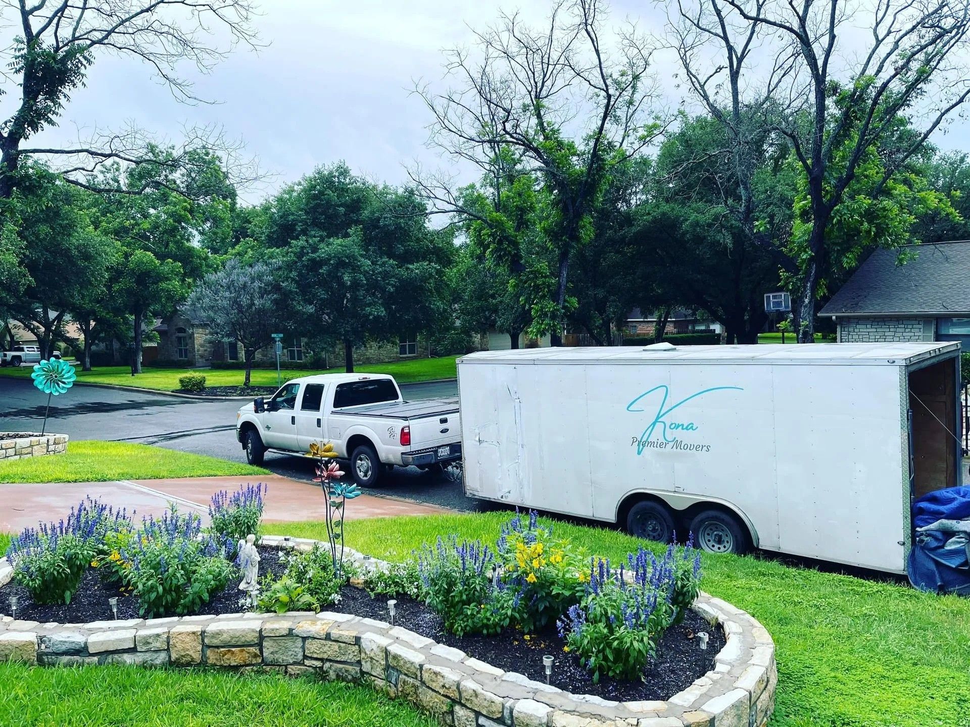 White pickup truck and trailer parked by a landscaped curb on a rainy residential street.