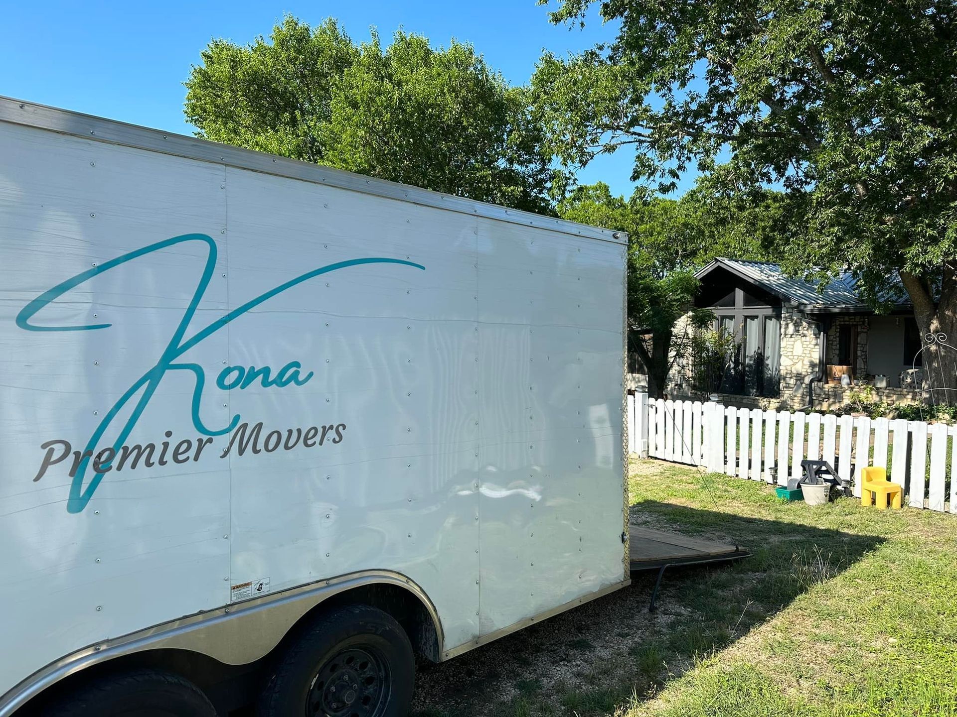 White moving trailer with “Zona Premier Movers” logo parked beside a white fence in a sunny yard