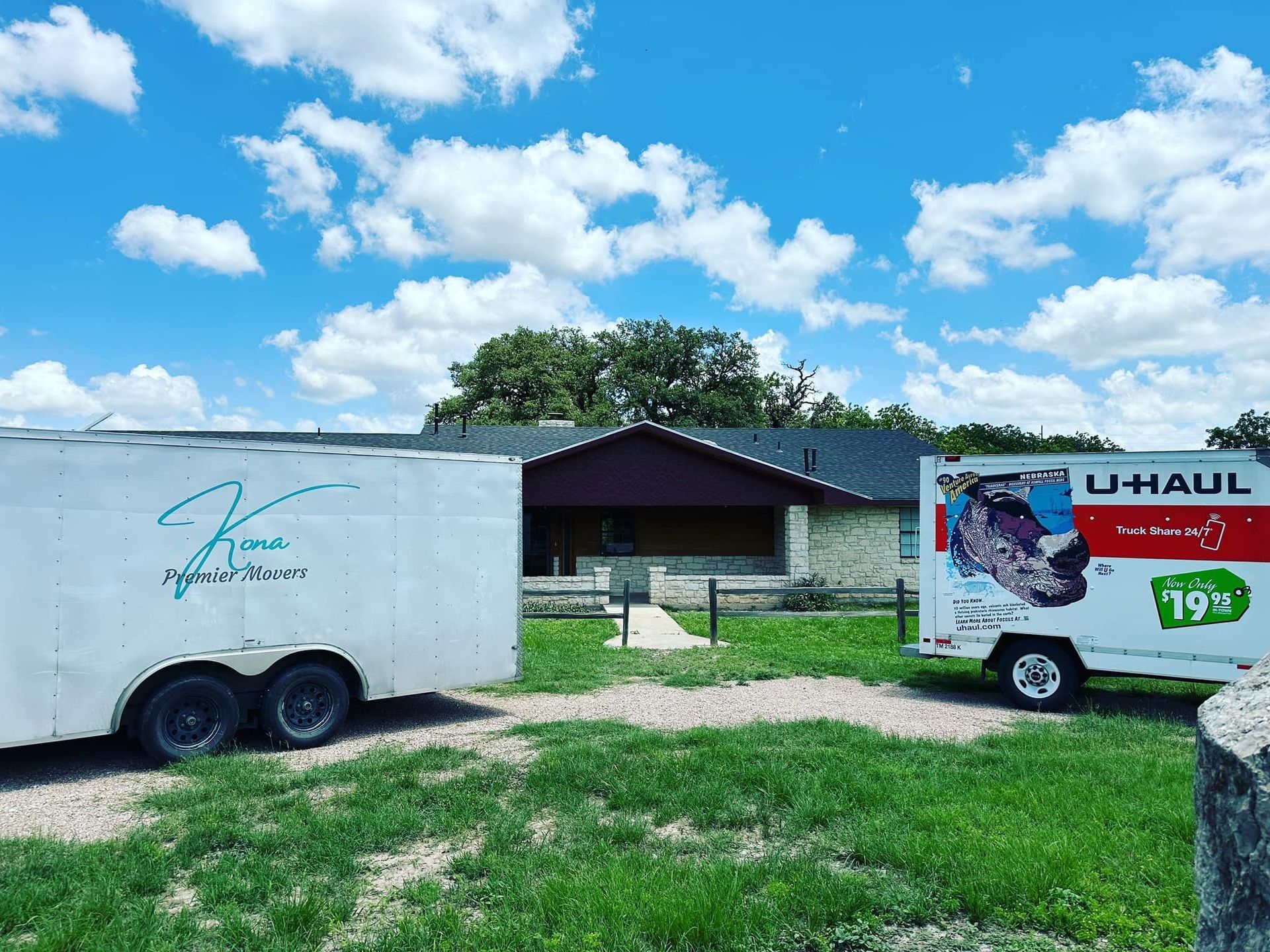 Two U-Haul trailers parked on a grassy lot near a small building under a bright blue sky.