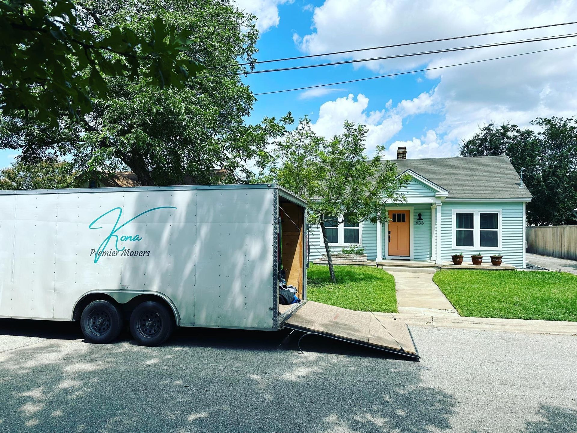 White moving trailer parked beside a small house, with a ramp down and trees overhead.