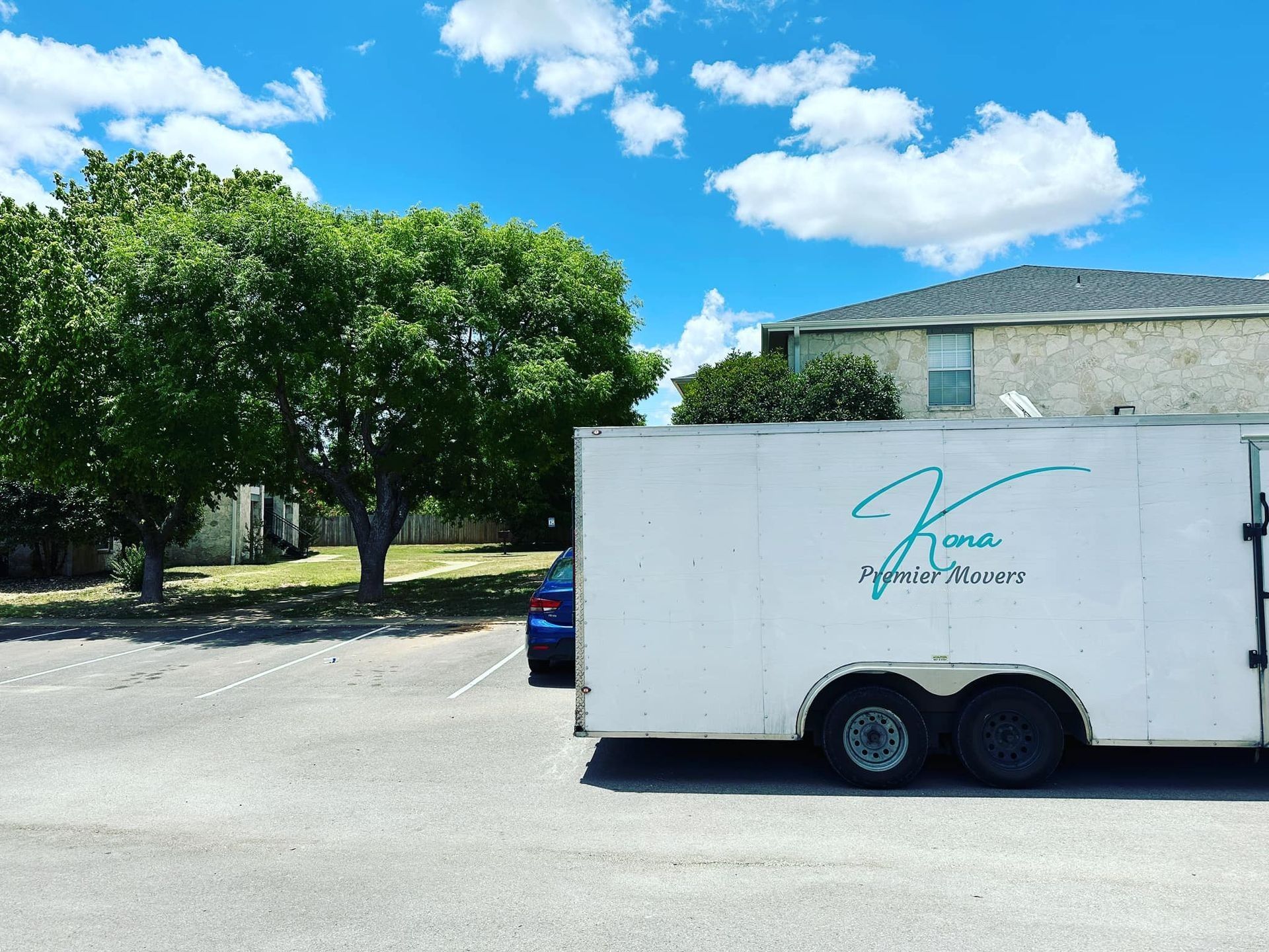 White trailer with teal logo parked by a building under a bright blue sky, beside a leafy tree.