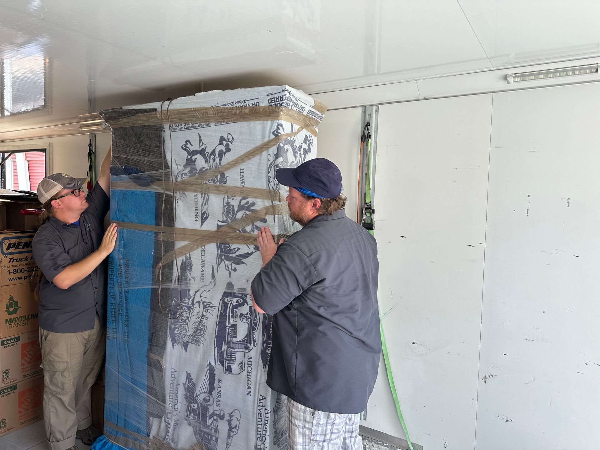 Two workers securing a large wrapped cabinet in a utility room with tape and straps.