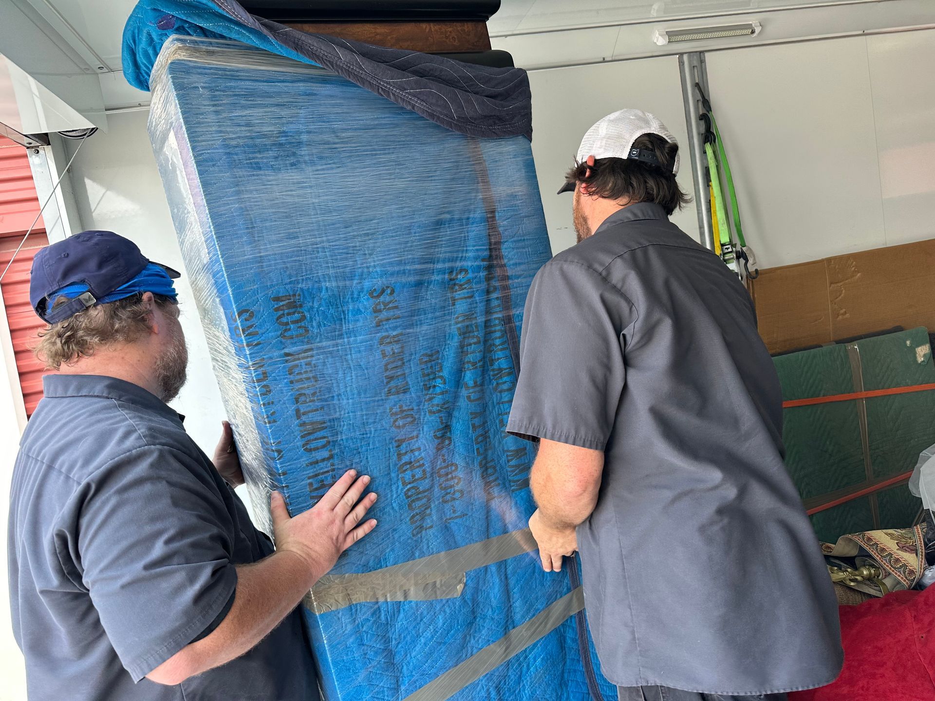 Two workers moving a large blue-wrapped item in a garage or storage area.