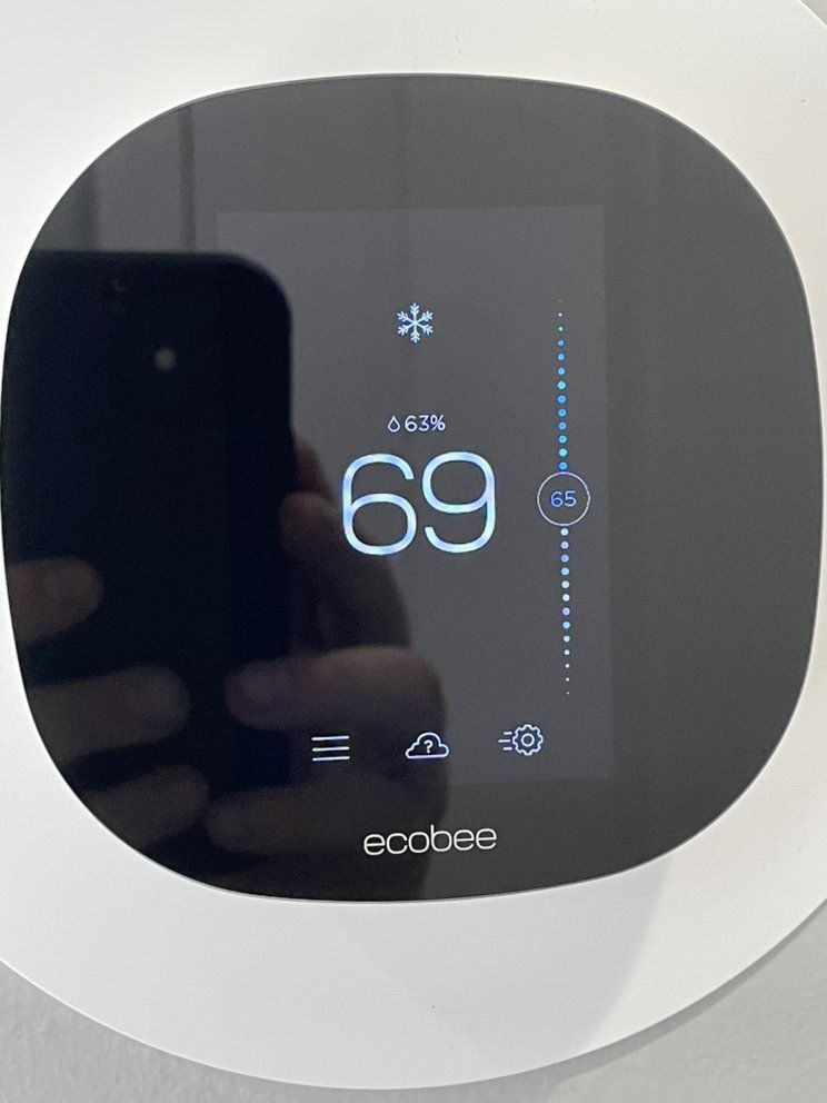 A person is holding a cell phone in front of a thermostat that says 69 degrees