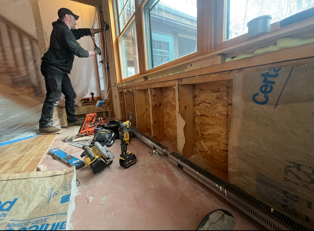 A person works on a wall near a window during construction. Tools and materials are on the floor.