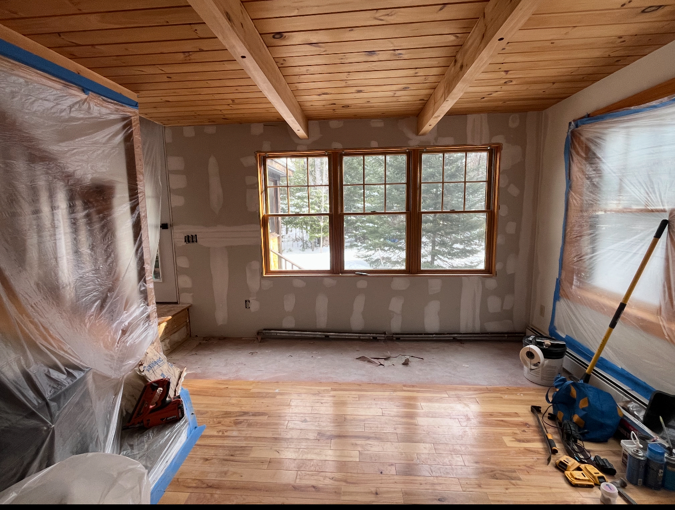 Room under renovation with exposed wood ceiling and trim, drywall walls, windows, and hardwood floors.
