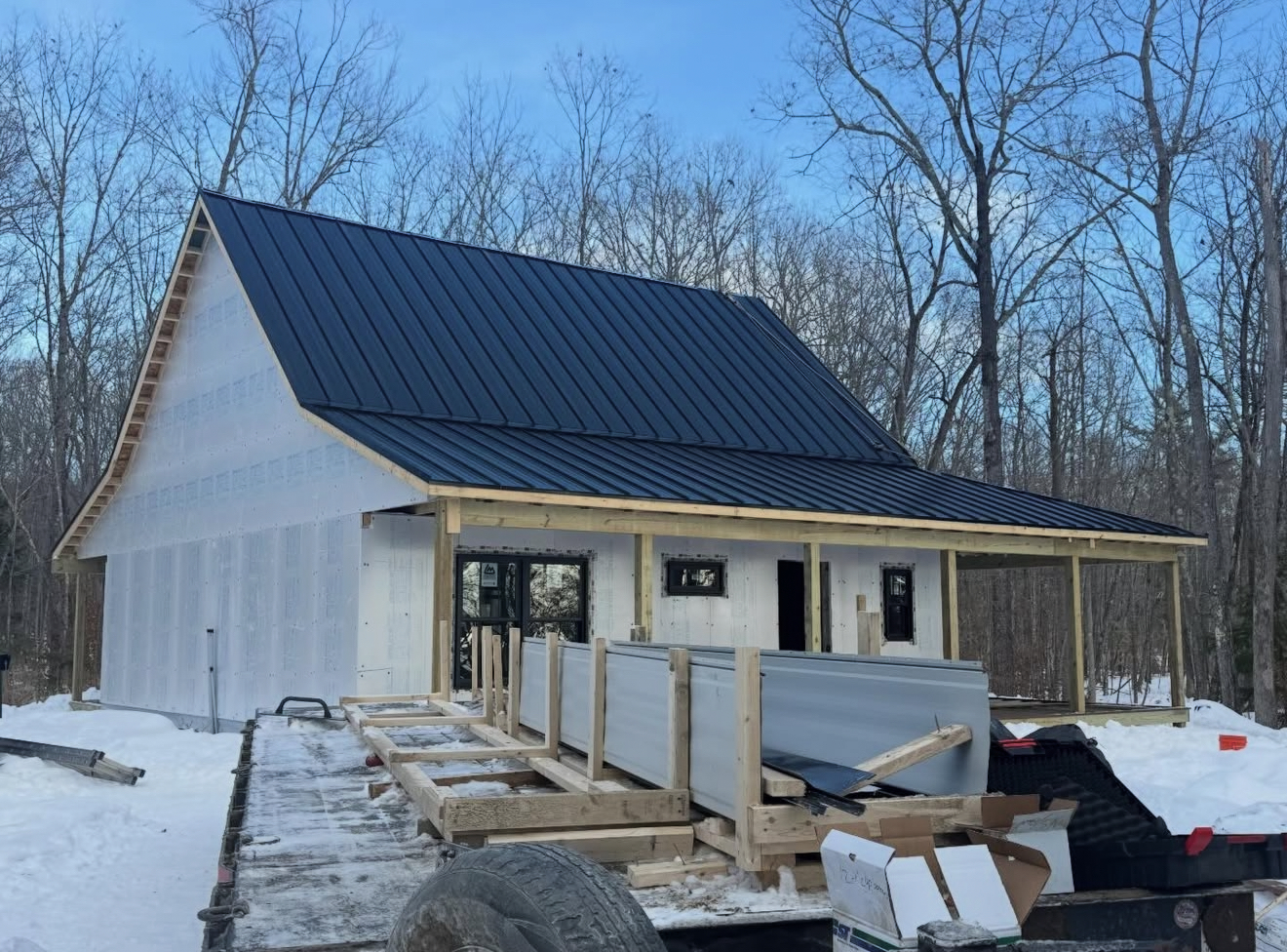 Cabin under construction with black roof and white siding, in a snowy, wooded area.