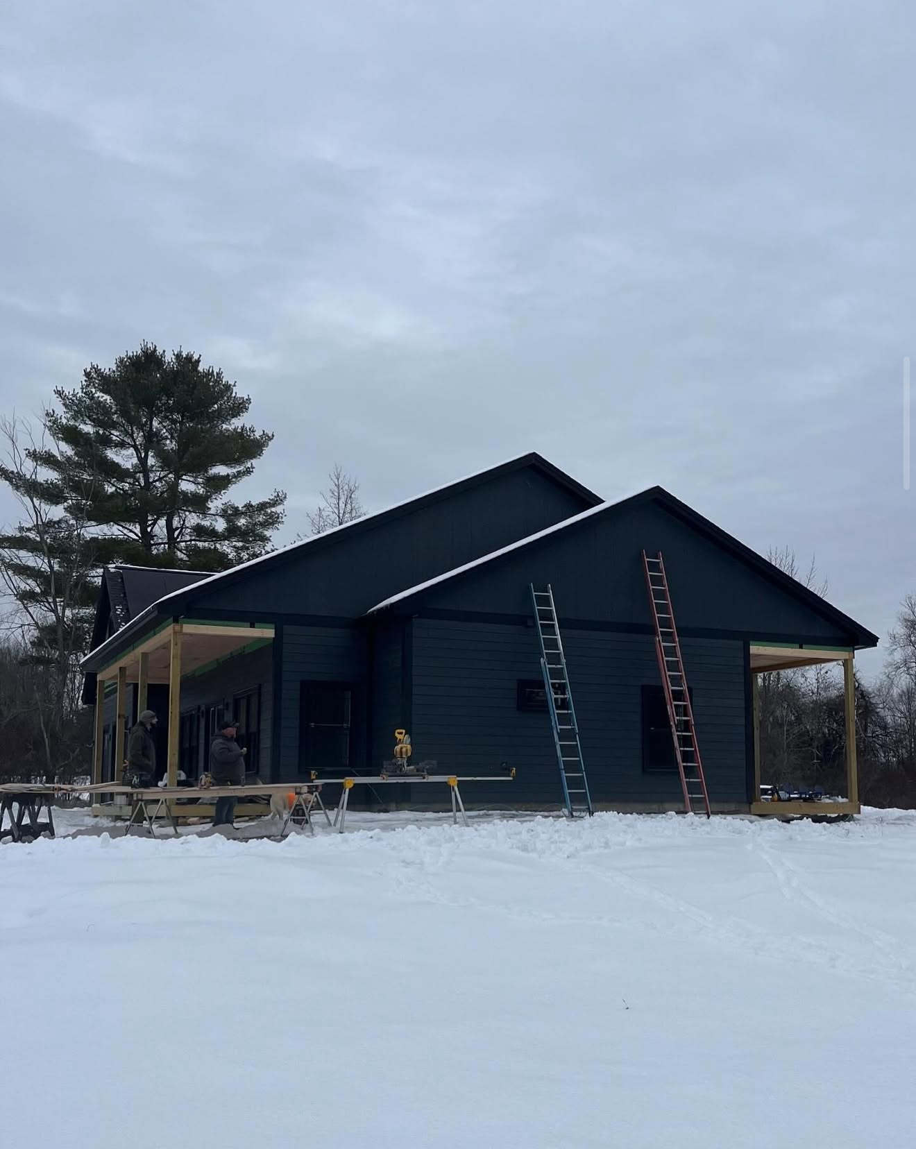 A house under construction, dark blue siding, wooden porch, snow-covered ground, ladders leaned against the wall.