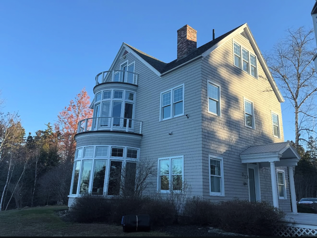 Two-story gray house with large windows, a curved balcony, and a brick chimney under a blue sky.