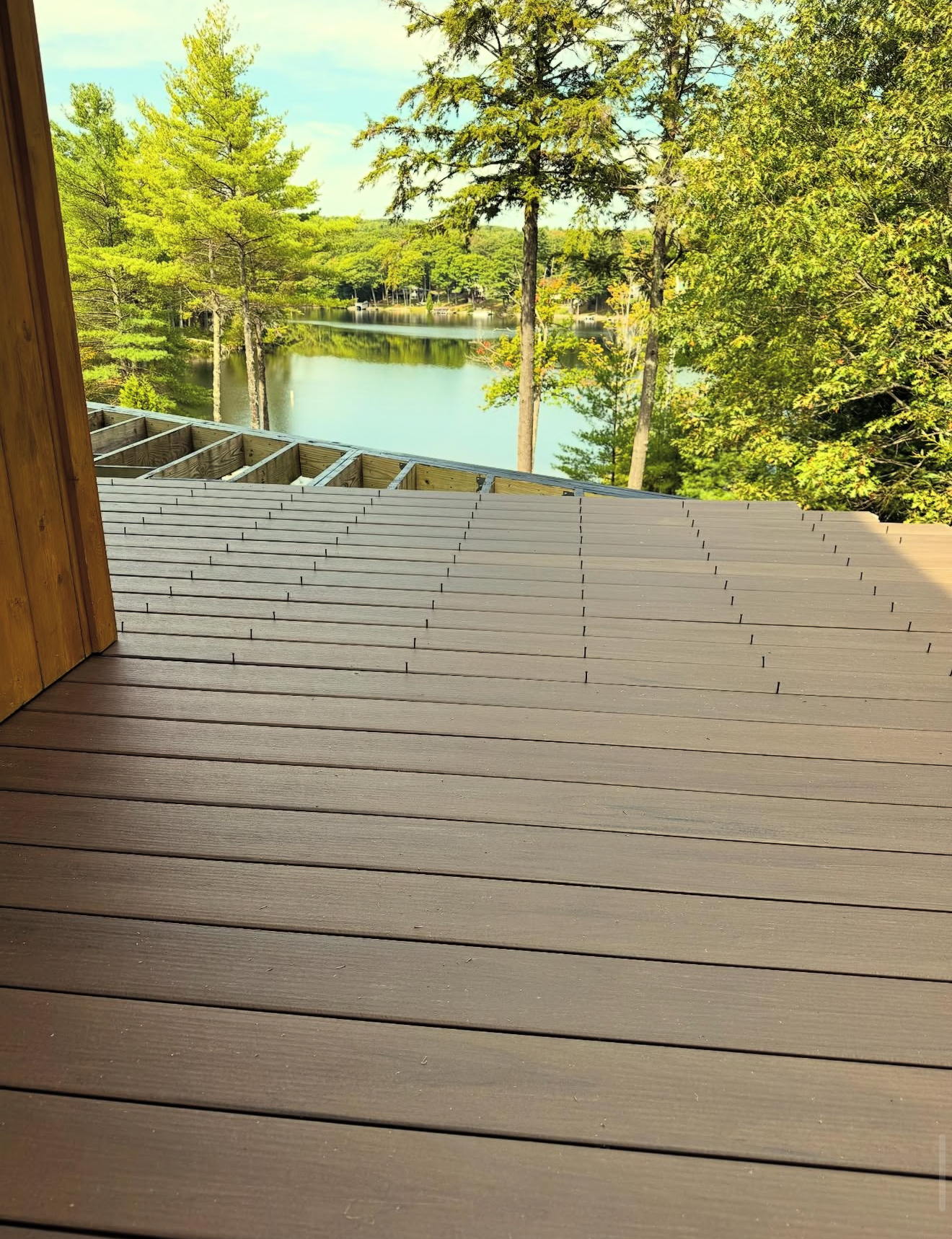 Wooden deck overlooking a lake, surrounded by trees. Brown decking, light blue water, and green foliage.