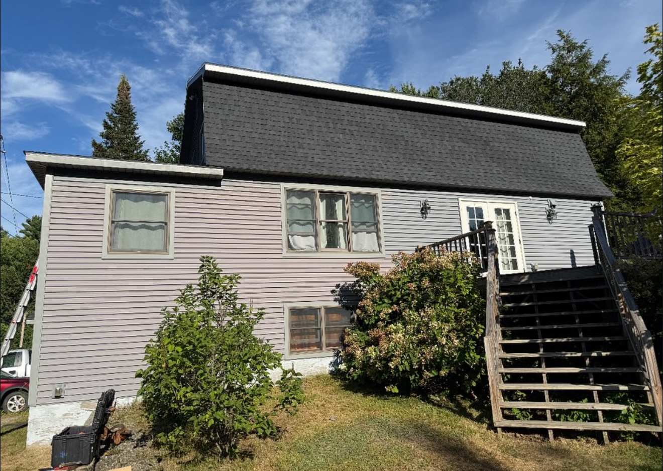 Two-story house with gray siding, black roof, wooden stairs, and overgrown landscaping.