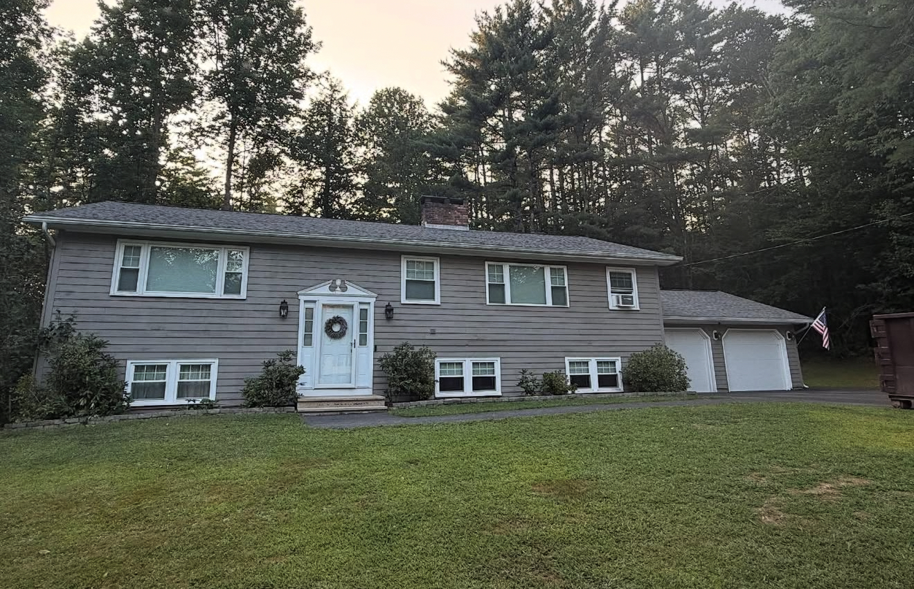 Gray house with white-trimmed windows and door, two-car garage, and green lawn, set against a backdrop of trees.