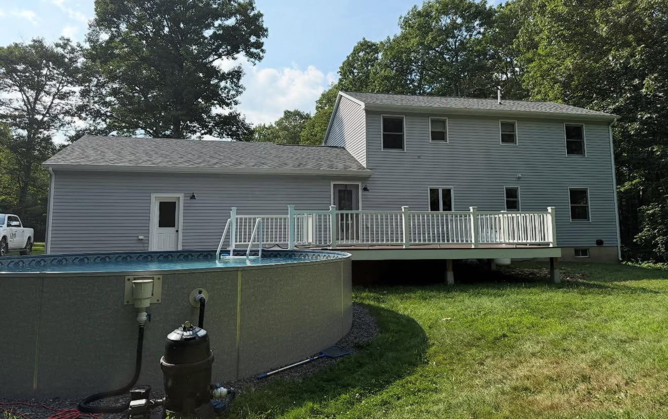 Backyard with above-ground pool, deck, and house with blue siding under a sunny sky.