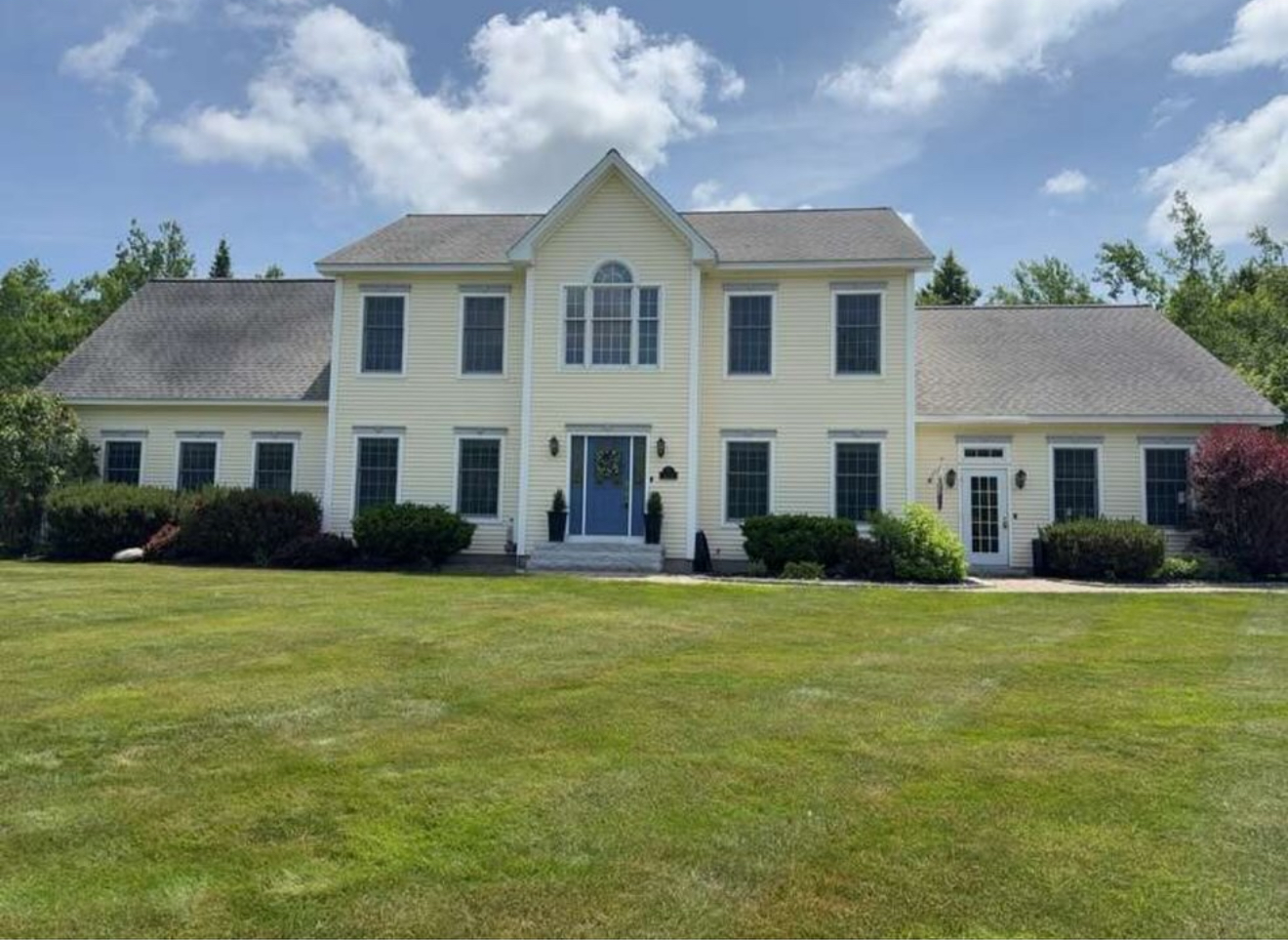 Yellow two-story house with blue door and large lawn, under a partly cloudy sky.