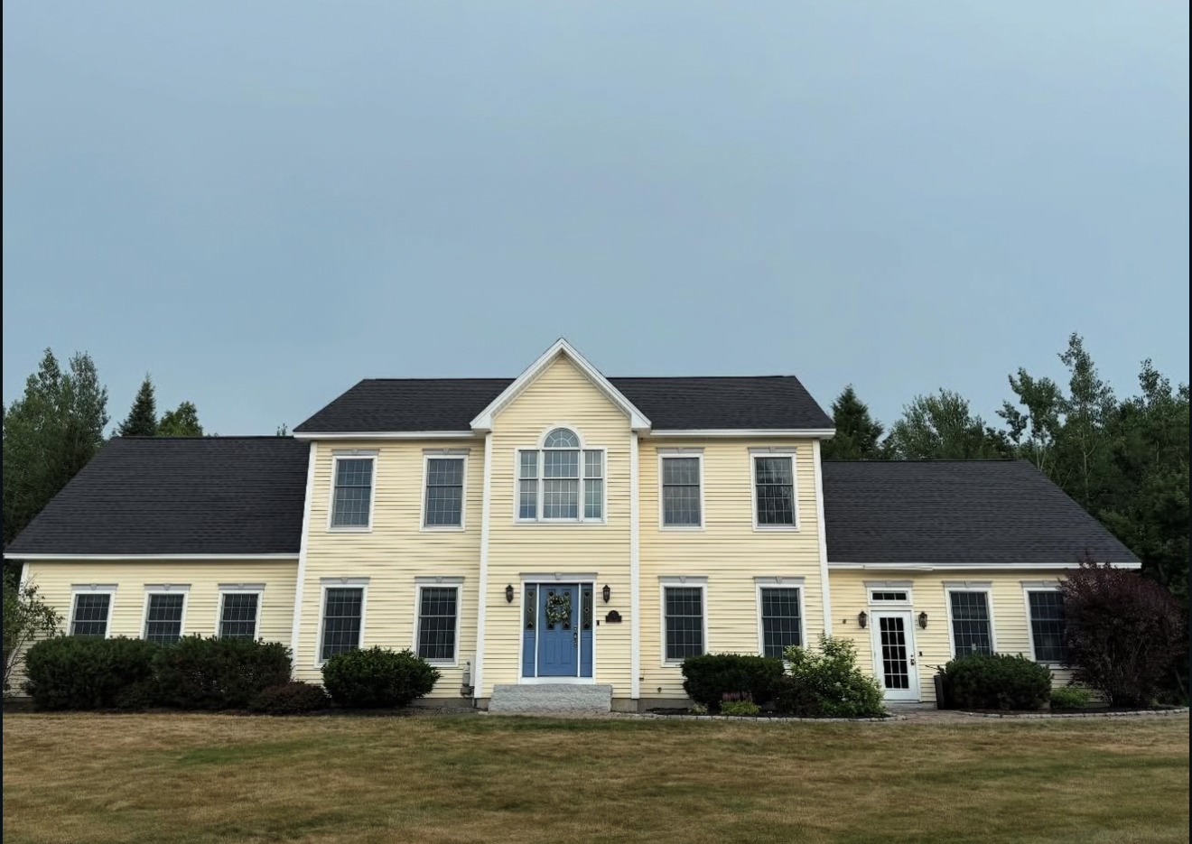 Large two-story yellow house with black roof and blue front door, set on a grassy lawn under a cloudy sky.