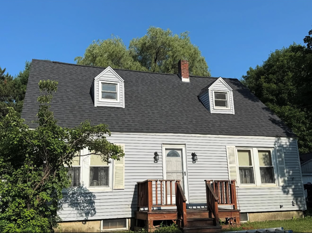 Weathered white house with black roof, two dormers, brown chimney, and small porch. Blue sky, green trees.