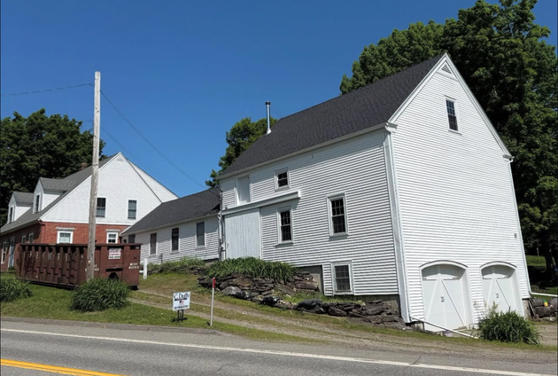 White clapboard building with black roof and two garage doors, set beside a road on a sunny day.
