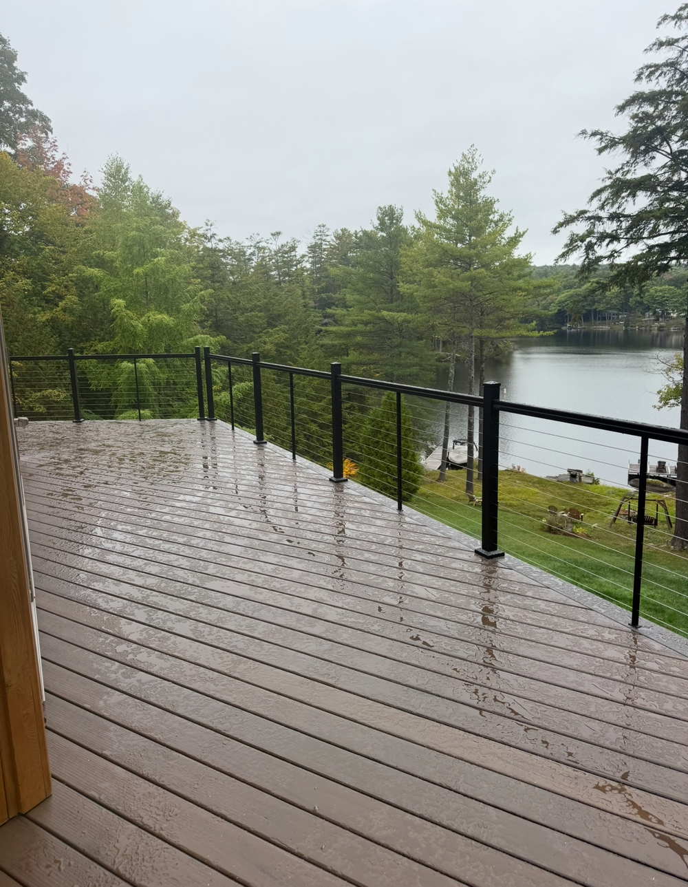 A wet, wood deck with a black railing overlooks a lake and green trees under a gray sky.