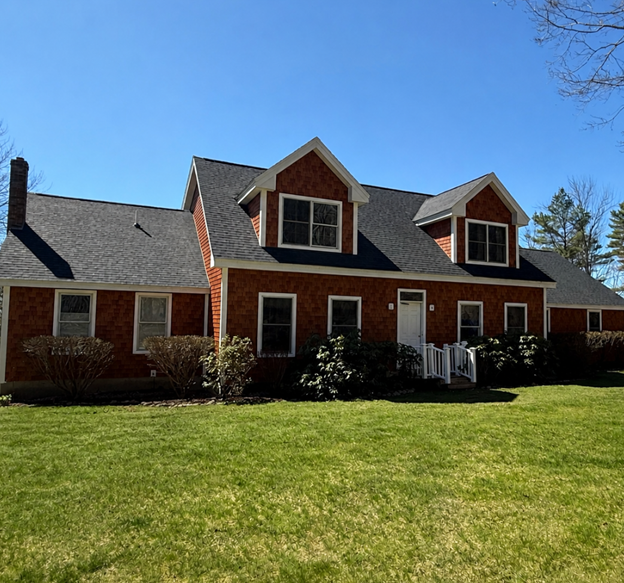 Two-story blue house with brown trim, wooden garage doors, and front porch, set in a wooded area.