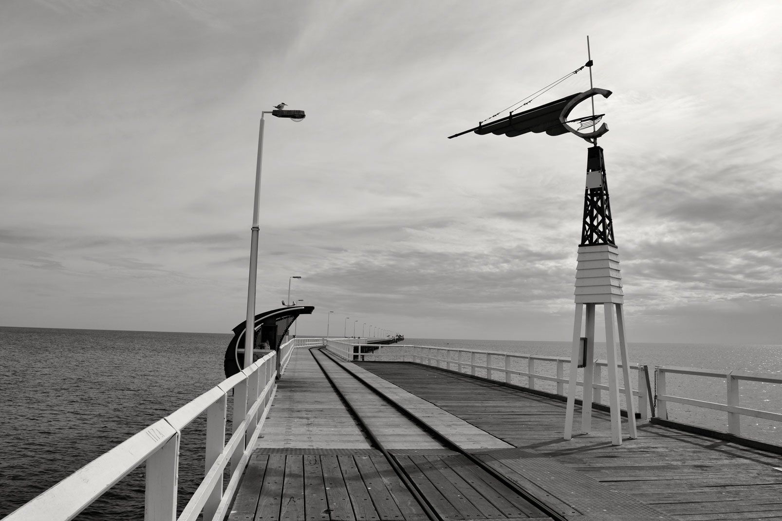 Black-and-white view of a pier with a crane-like structure over calm water under a cloudy sky