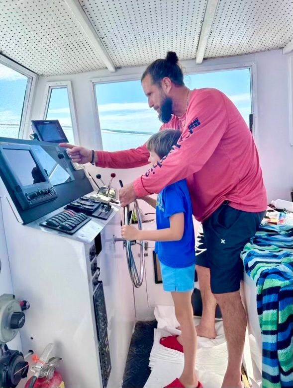 Man and child using a touchscreen console in a bright control room with windows and monitors.