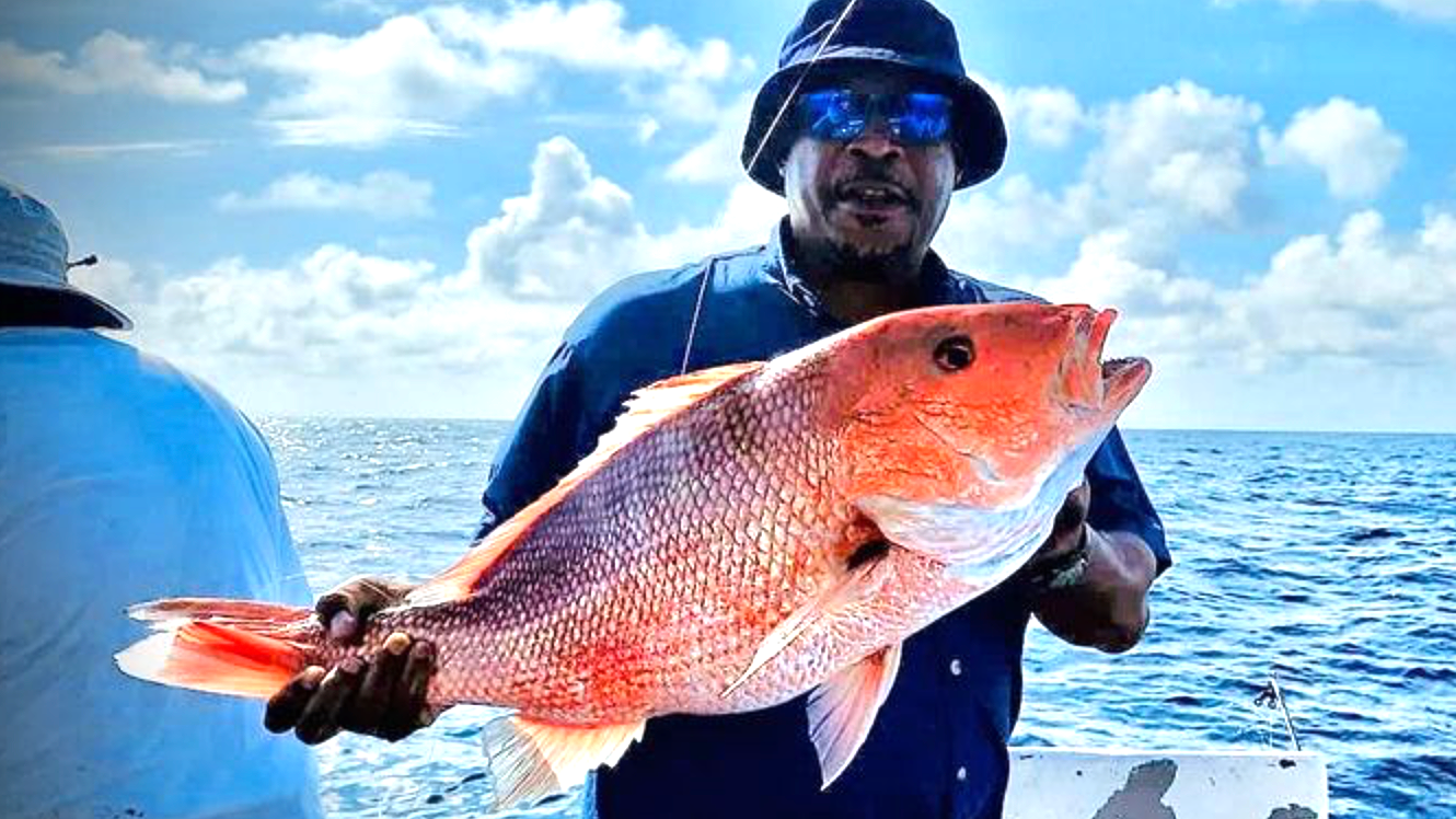 A person wearing a sun hat and sunglasses holds a large red snapper fish on a boat against a blue sky and ocean.