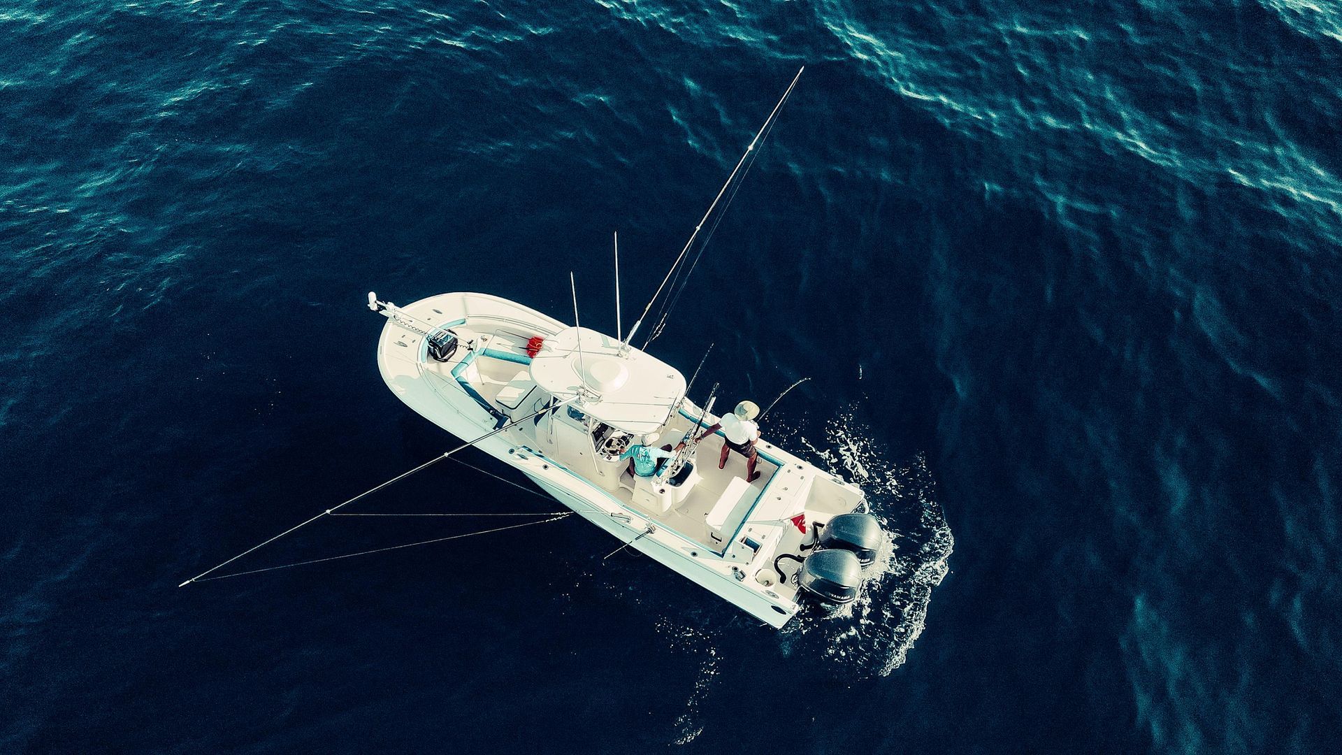 A white sailboat in dark blue ocean, seen from above with sails furled and crew on deck