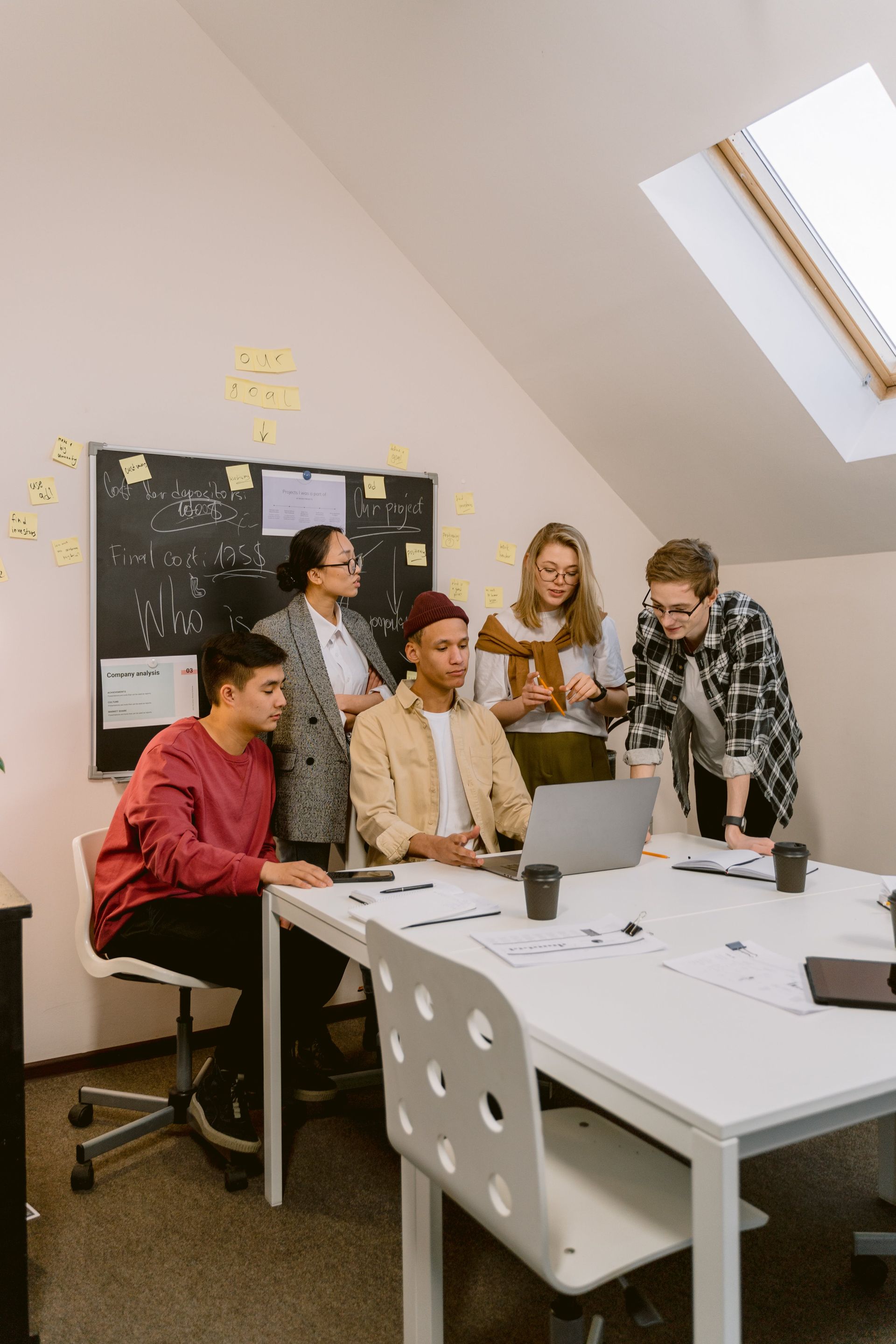 A group of people collaborating around a laptop at a table in a bright office with a blackboard and sticky notes.