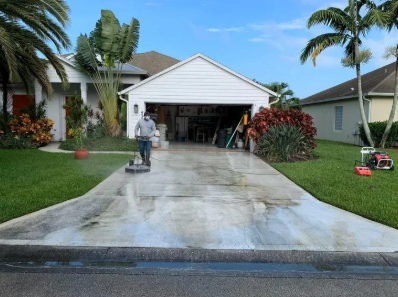 Person in yellow boots pressure washing a brick patio beside green shrubs and pink flowers