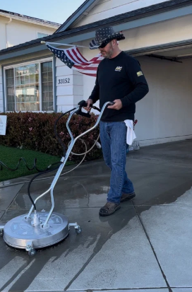 Person in yellow boots pressure washing a brick patio beside green shrubs and pink flowers