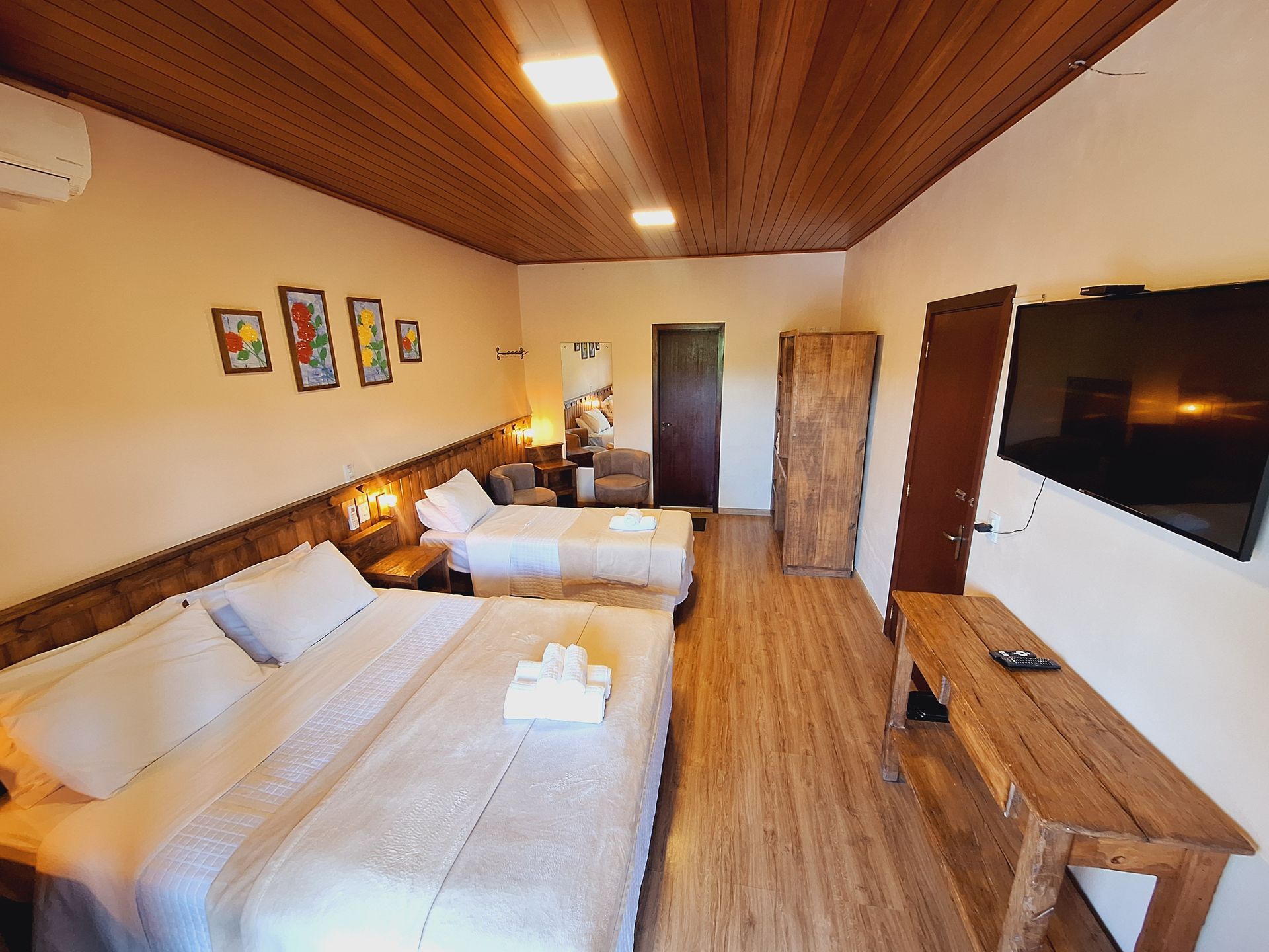 Um quarto de hotel com duas camas, teto e piso de madeira, obras de arte nas paredes, uma TV de tela plana e uma mesa de console rústica de madeira.