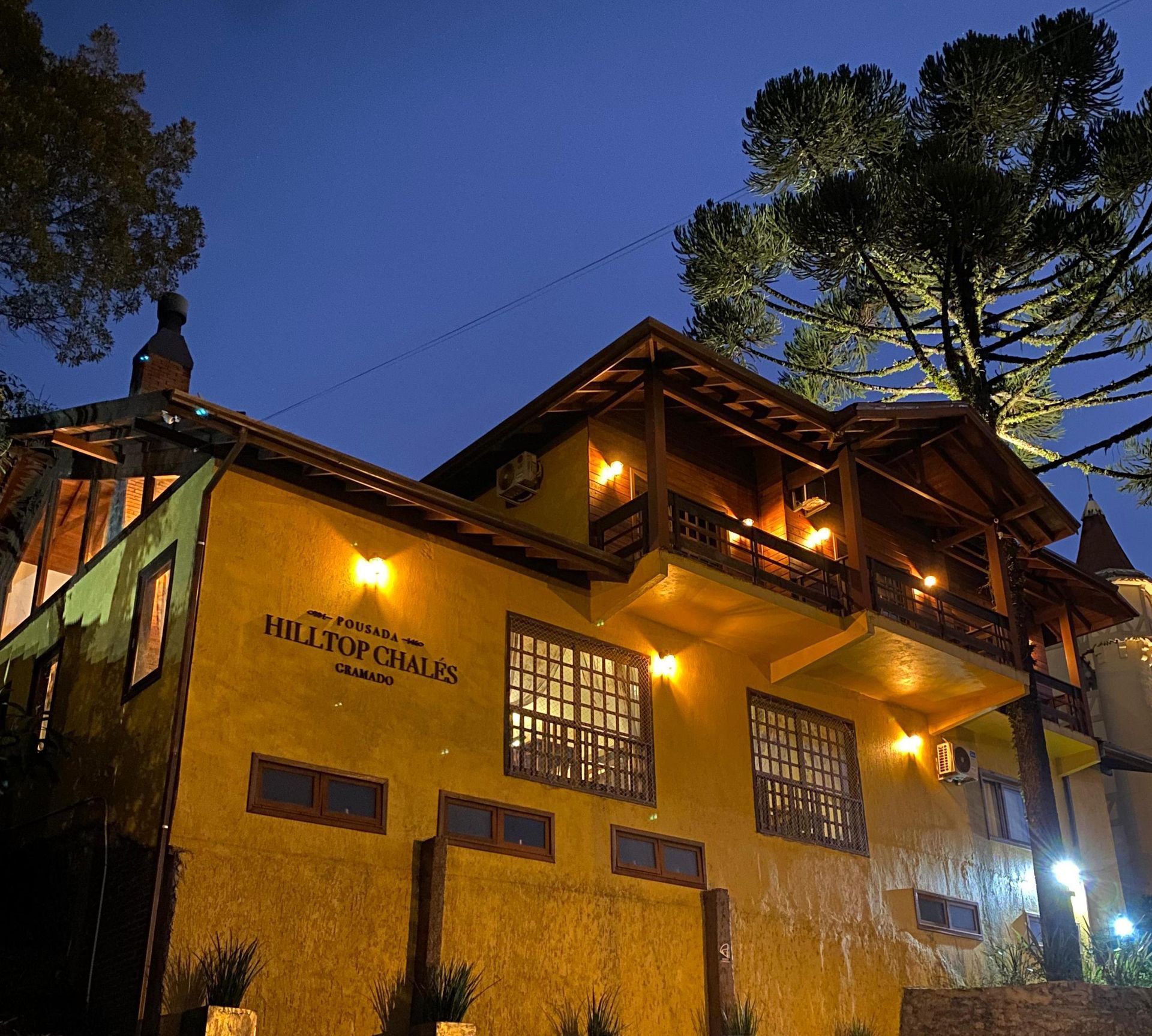 Um prédio rústico amarelo de vários andares à noite, com varandas iluminadas, detalhes em madeira e uma grande árvore contra um céu escuro.