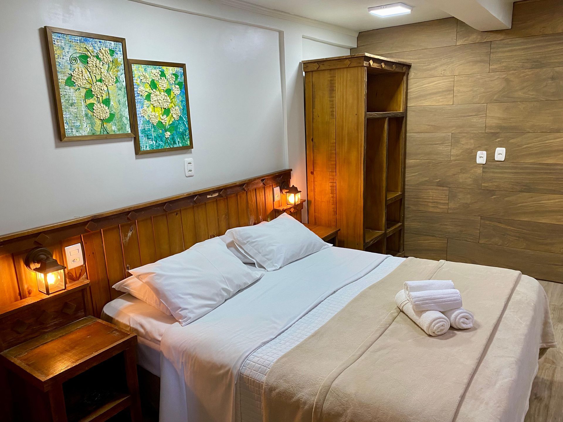 Um quarto de hotel com uma cama queen-size com lençóis brancos, cabeceira de madeira, mesas de cabeceira e duas pinturas florais.