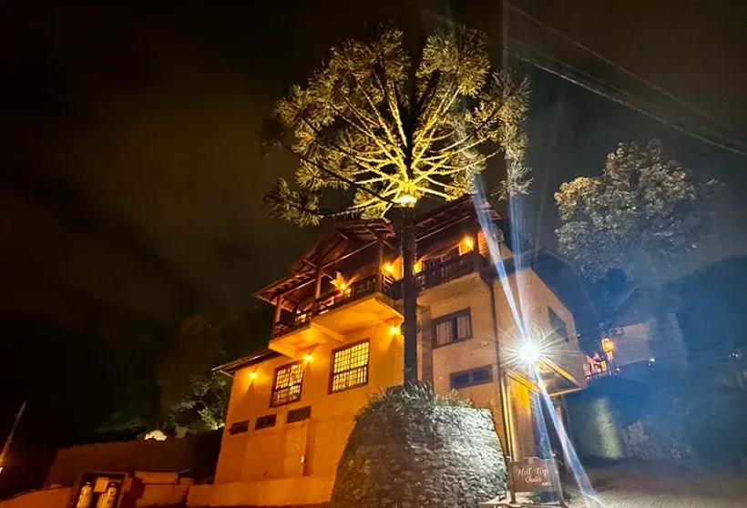 Um chalé iluminado de dois andares à noite, com uma imponente araucária iluminada por holofotes em primeiro plano.