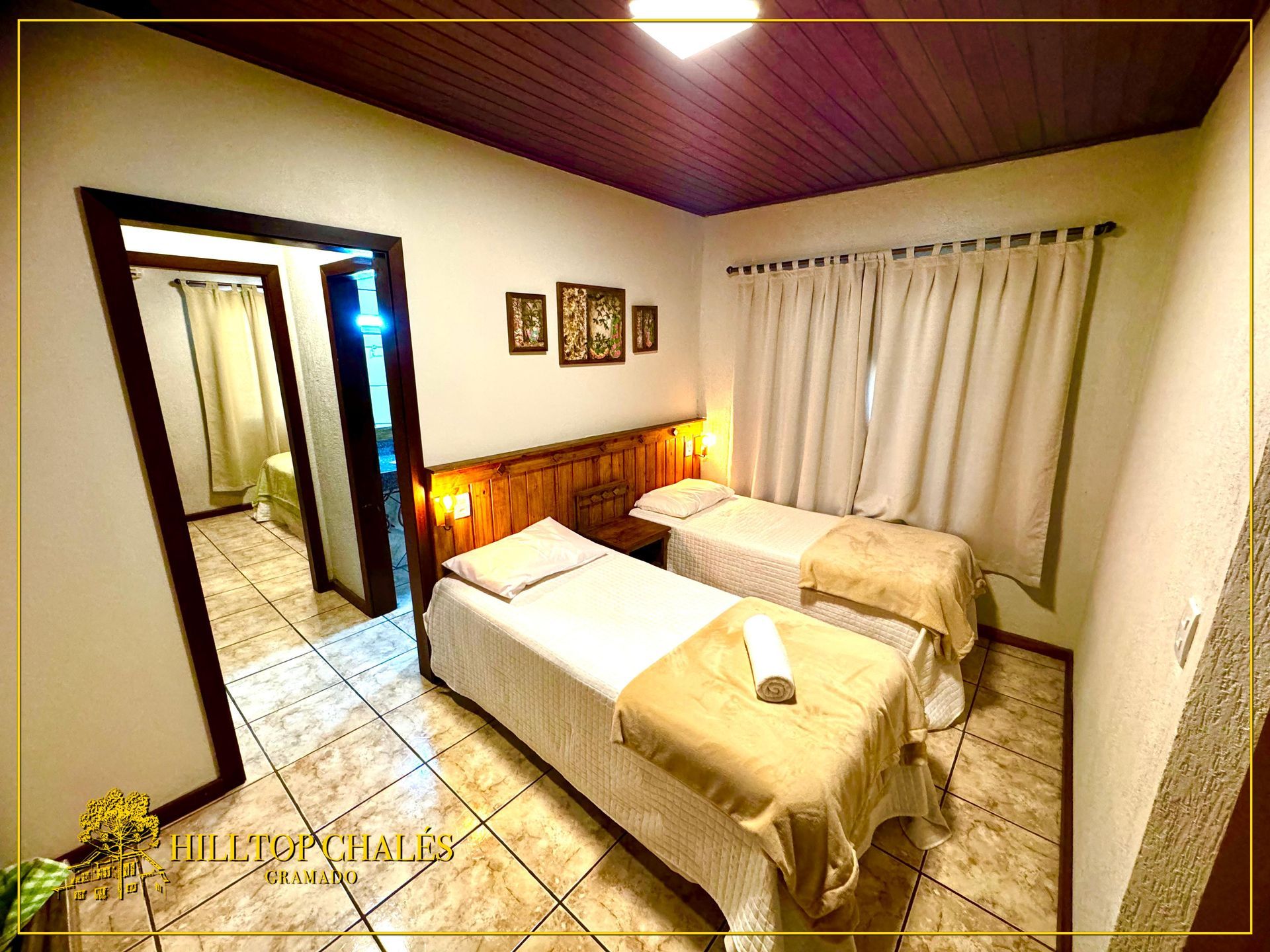 Um quarto aconchegante com duas camas de solteiro, teto com painéis de madeira, piso de azulejo e uma porta que dá para outro cômodo.