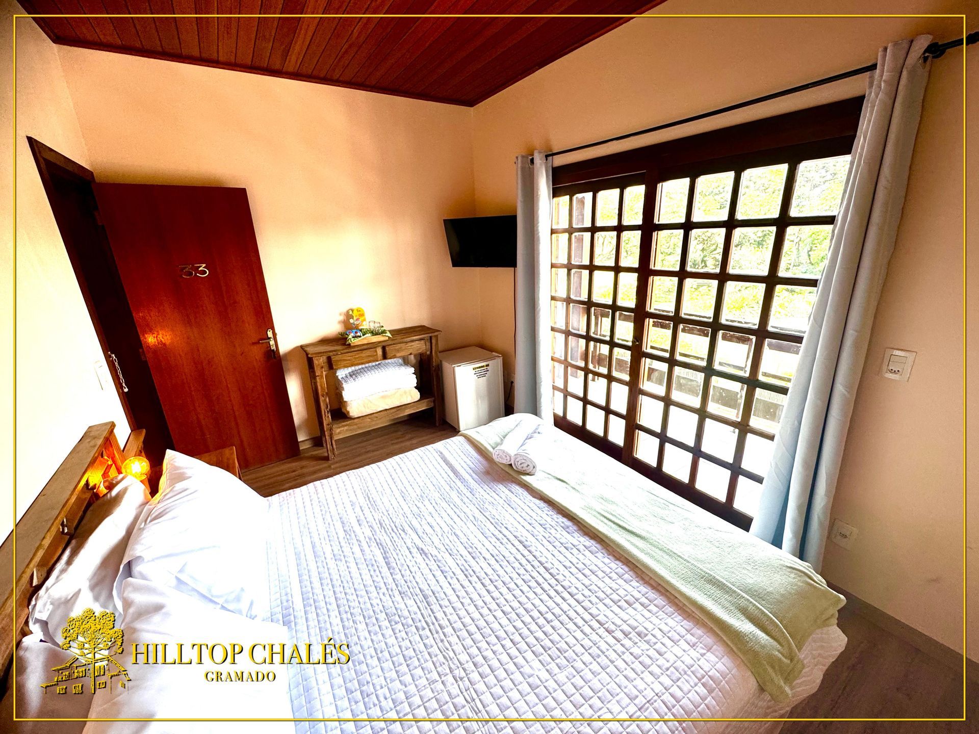 Um quarto aconchegante com uma cama bem arrumada, teto de madeira, porta de madeira escura e porta de vidro deslizante com vista para a vegetação.