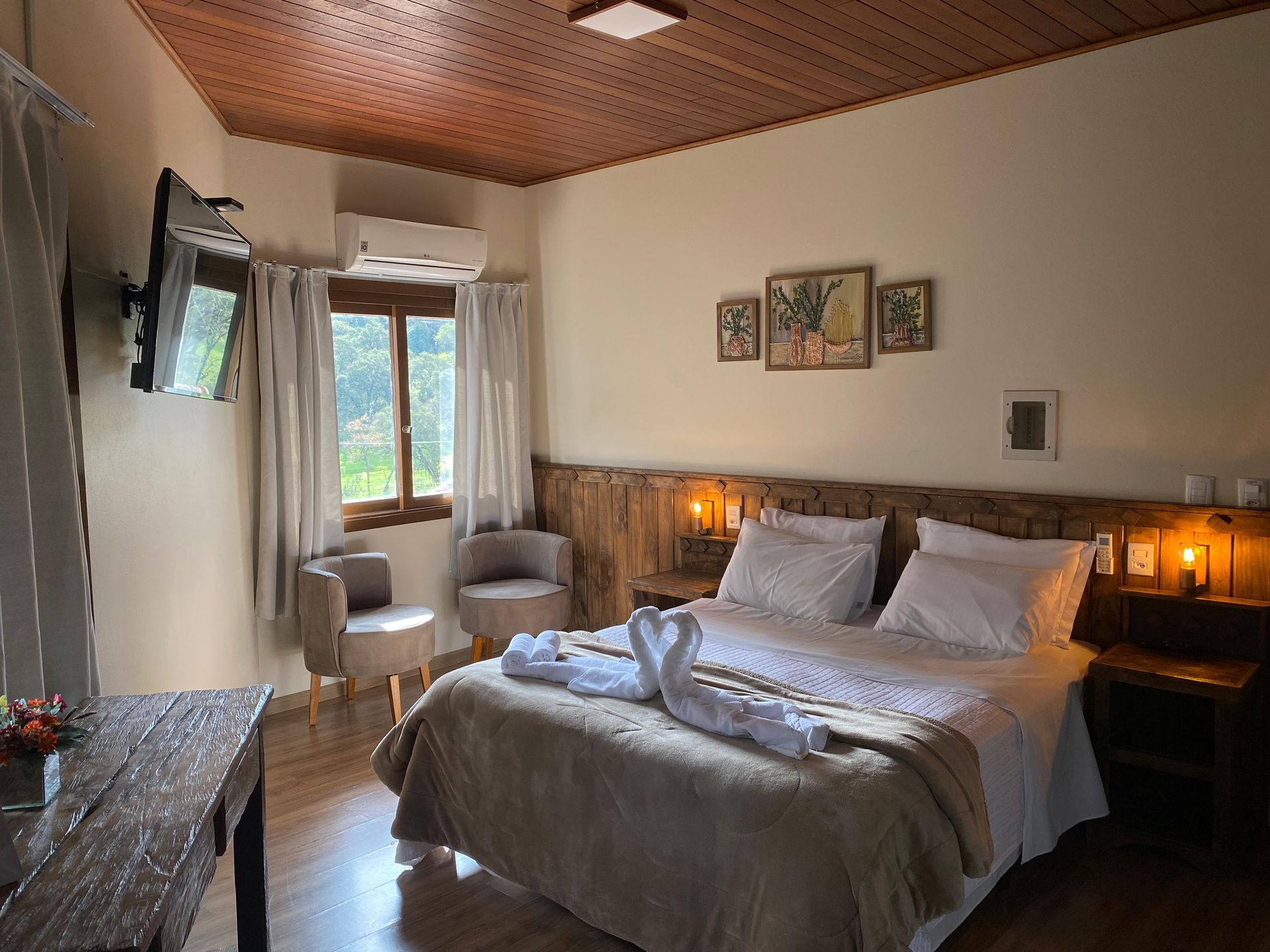 Um quarto de hotel aconchegante com uma cama queen-size, cabeceira de madeira, duas poltronas, uma TV fixada na parede e iluminação ambiente acolhedora.