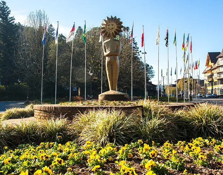 Uma estátua com cabeça em forma de sol ergue-se num canteiro circular, rodeada por um semicírculo de bandeiras hasteadas em mastros altos.