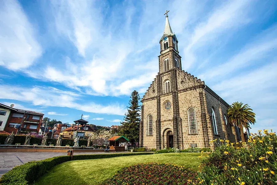 Uma igreja histórica de pedra com uma torre alta ergue-se ao lado de um jardim sob um céu azul brilhante com nuvens esparsas.